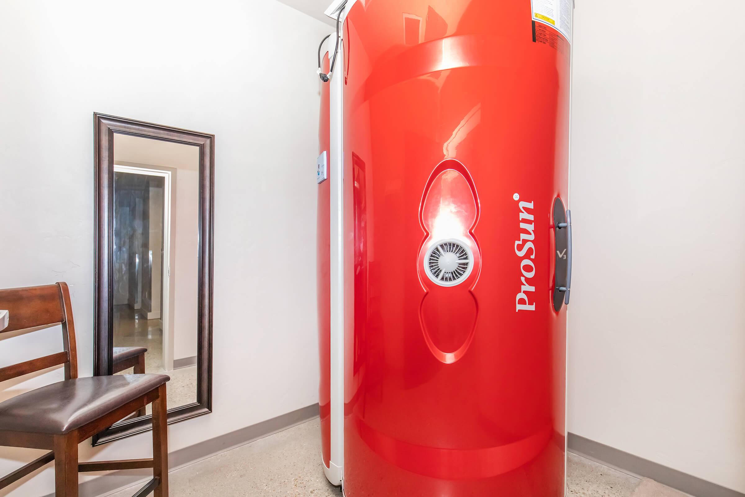 A bright red tanning bed labeled "ProSun" is positioned in a room with light-colored walls. There's a wooden chair nearby and a full-length mirror reflecting the room. The setting suggests a small tanning salon or personal tanning space.