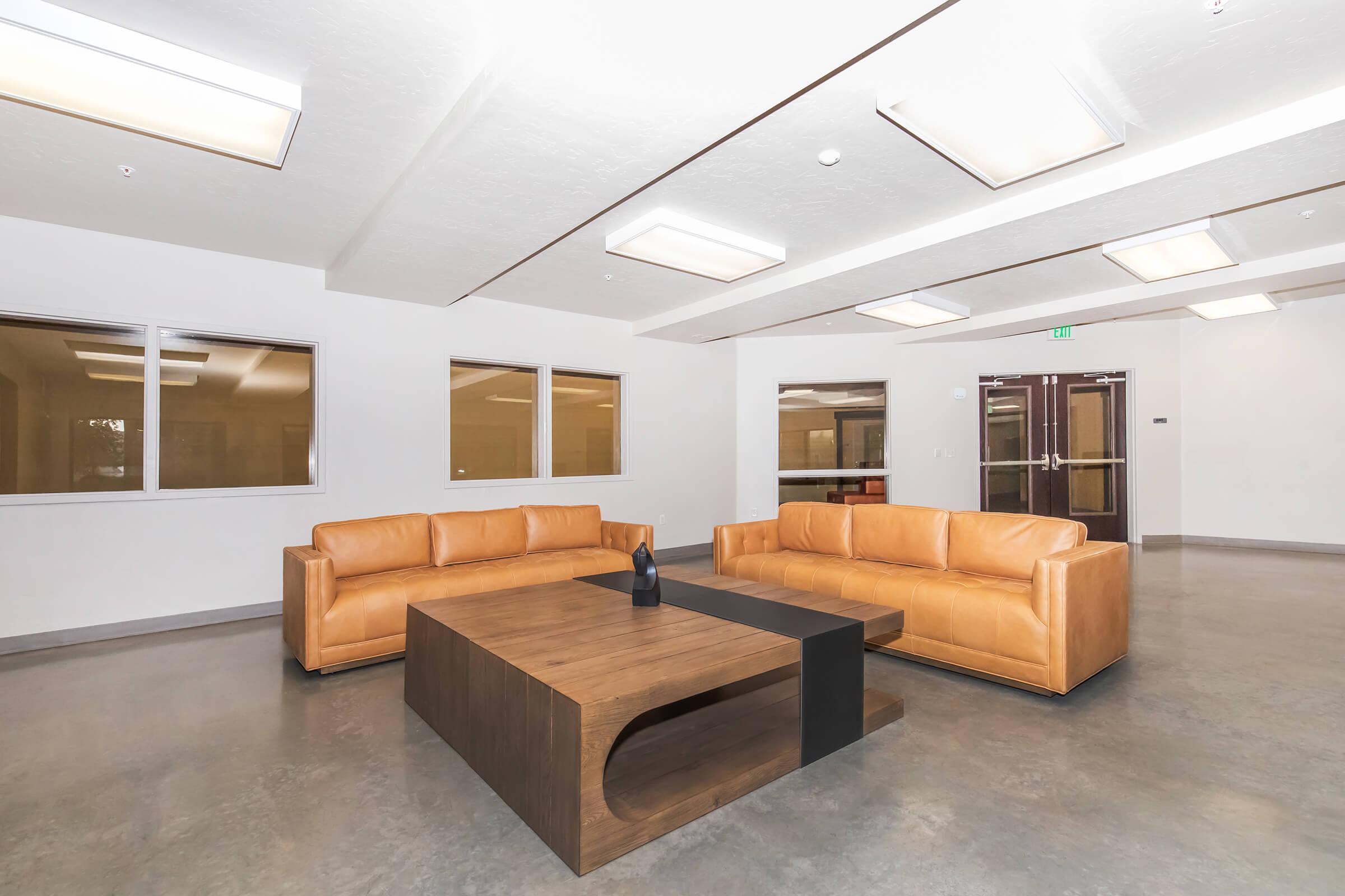 A modern lounge area featuring two tan leather sofas arranged around a wooden coffee table. The space has plain white walls, large windows allowing natural light, and a sleek concrete floor. There is a minimalist vase on the table, enhancing the contemporary aesthetic of the room.