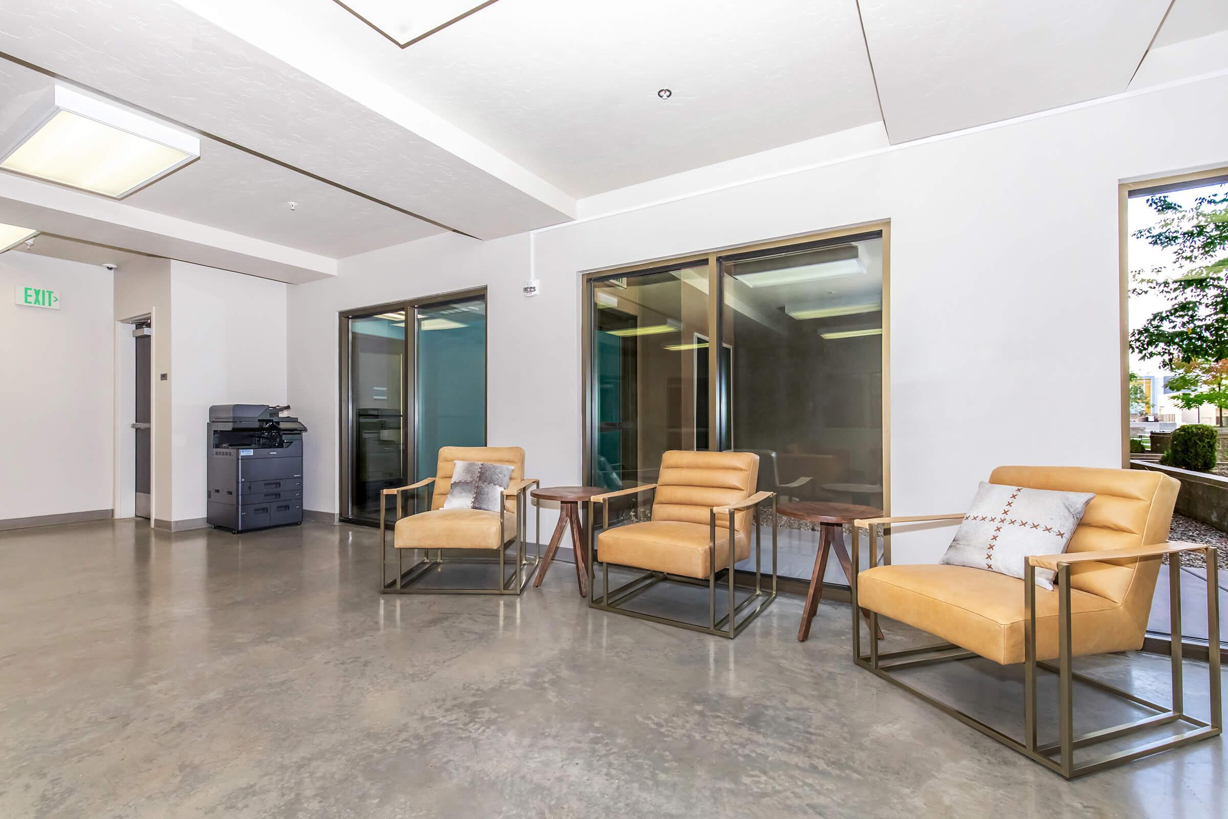 A modern waiting area featuring two stylish tan armchairs with decorative pillows, two small side tables, and a printer setup. Large windows allow natural light to fill the space, highlighting the polished concrete floor and neutral wall colors. The environment appears clean and inviting.