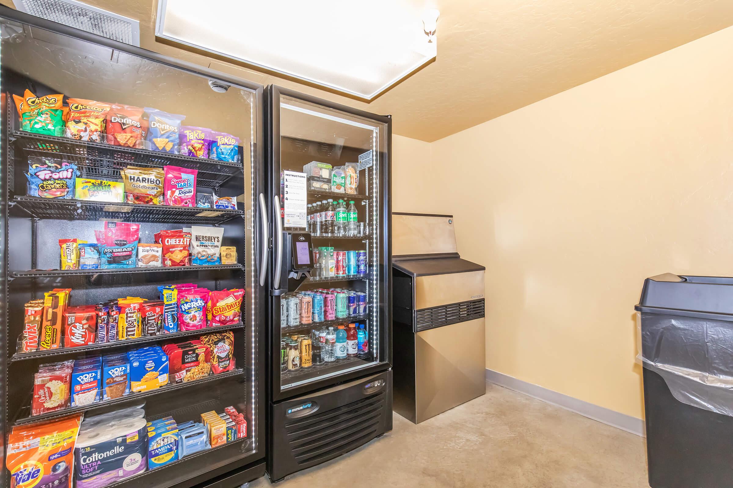 A well-stocked pantry with vending machines on the left displaying various snacks and beverages. On the right, there's an ice machine and a trash bin. The room has a light, neutral color scheme and bright overhead lighting.