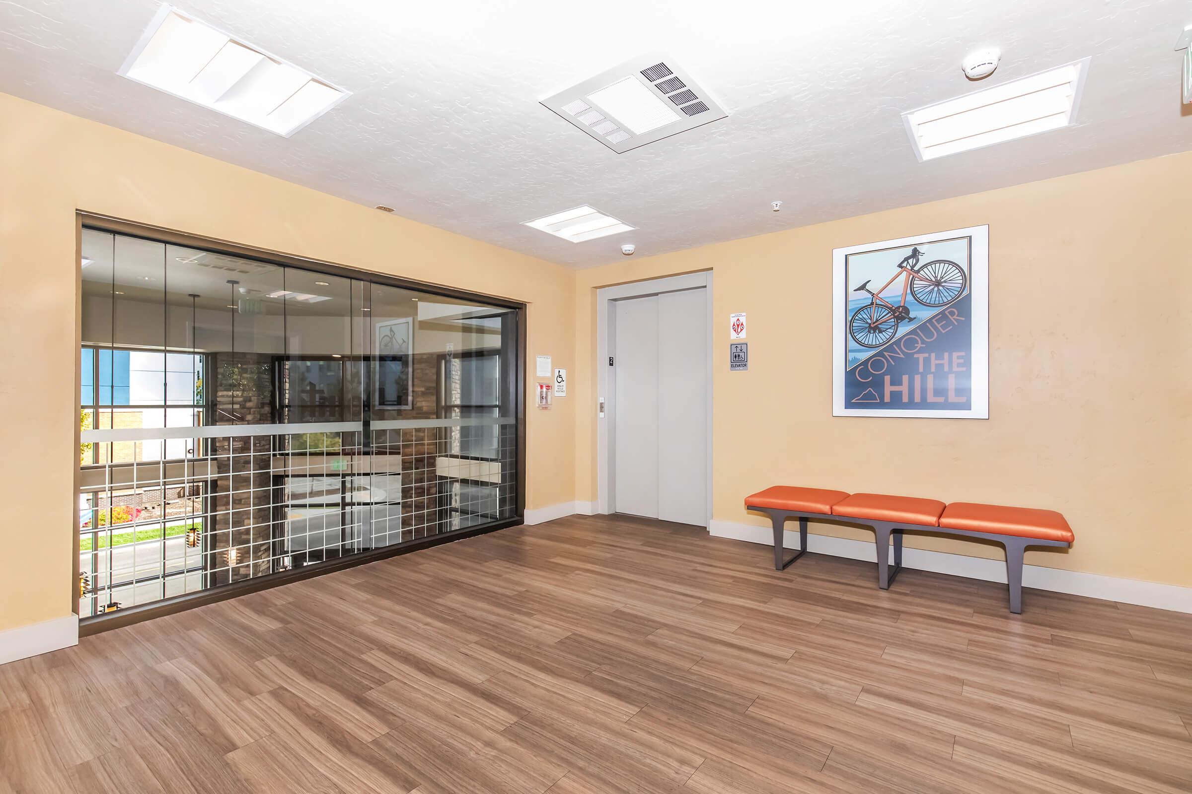 A spacious interior lobby with a warm orange wall color. There is a glass wall leading to an elevator and a framed poster featuring a bicycle with the text "CONQUER THE HILL." Two orange benches are positioned along the wall, and the floor features wood-like laminate tiles. Bright overhead lights illuminate the area.