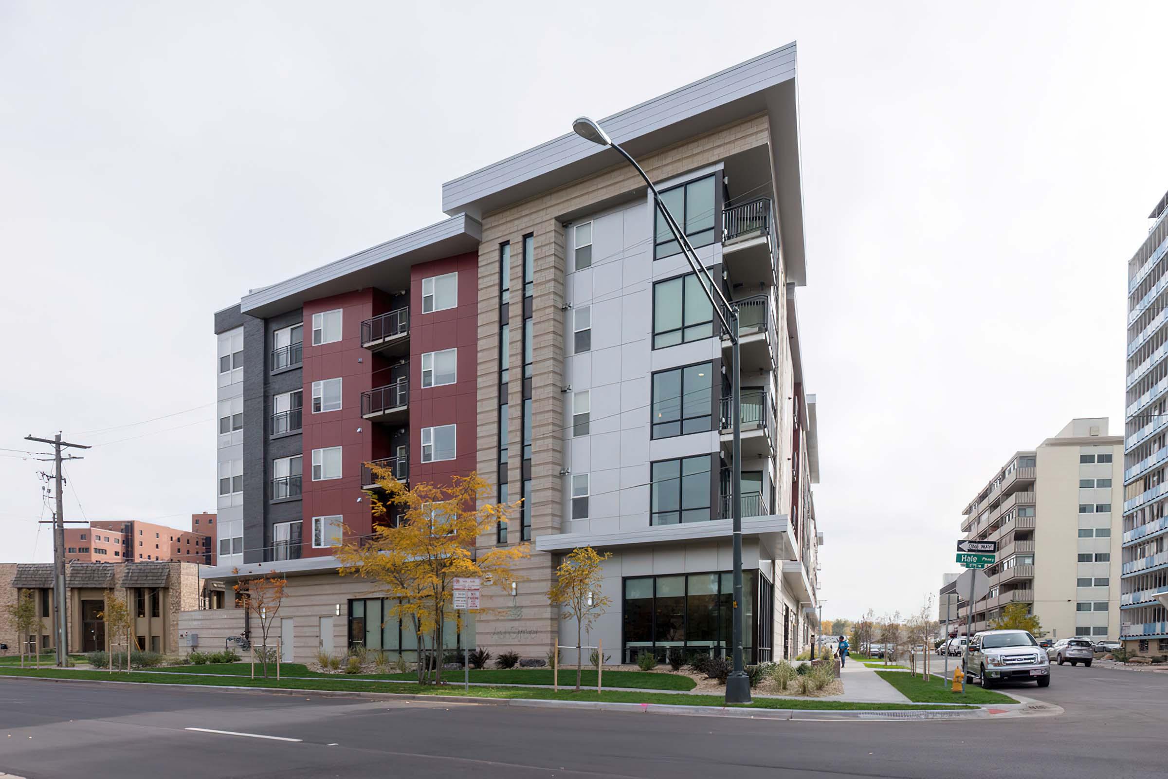Modern multi-story building featuring a mix of red and white exterior panels, large windows, and balconies. Street-level landscaping with small trees. Nearby, additional buildings and street signs are visible, with a cloudy sky in the background.