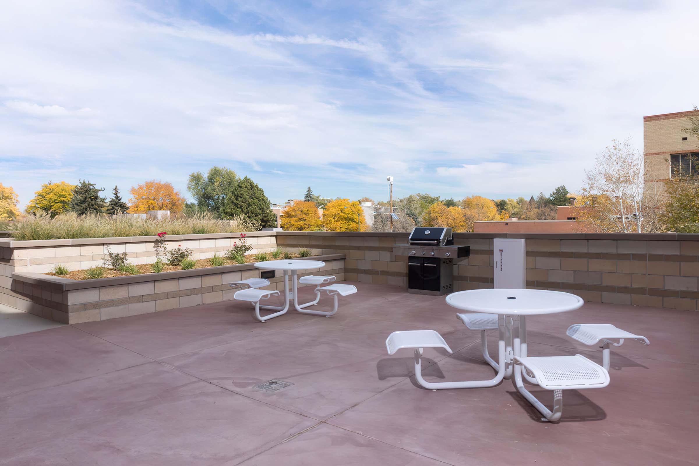A rooftop terrace featuring two white circular tables with attached seating, a grill in the background, and landscaped areas with trees displaying autumn foliage. The sky is partly cloudy, creating a bright and inviting outdoor space.