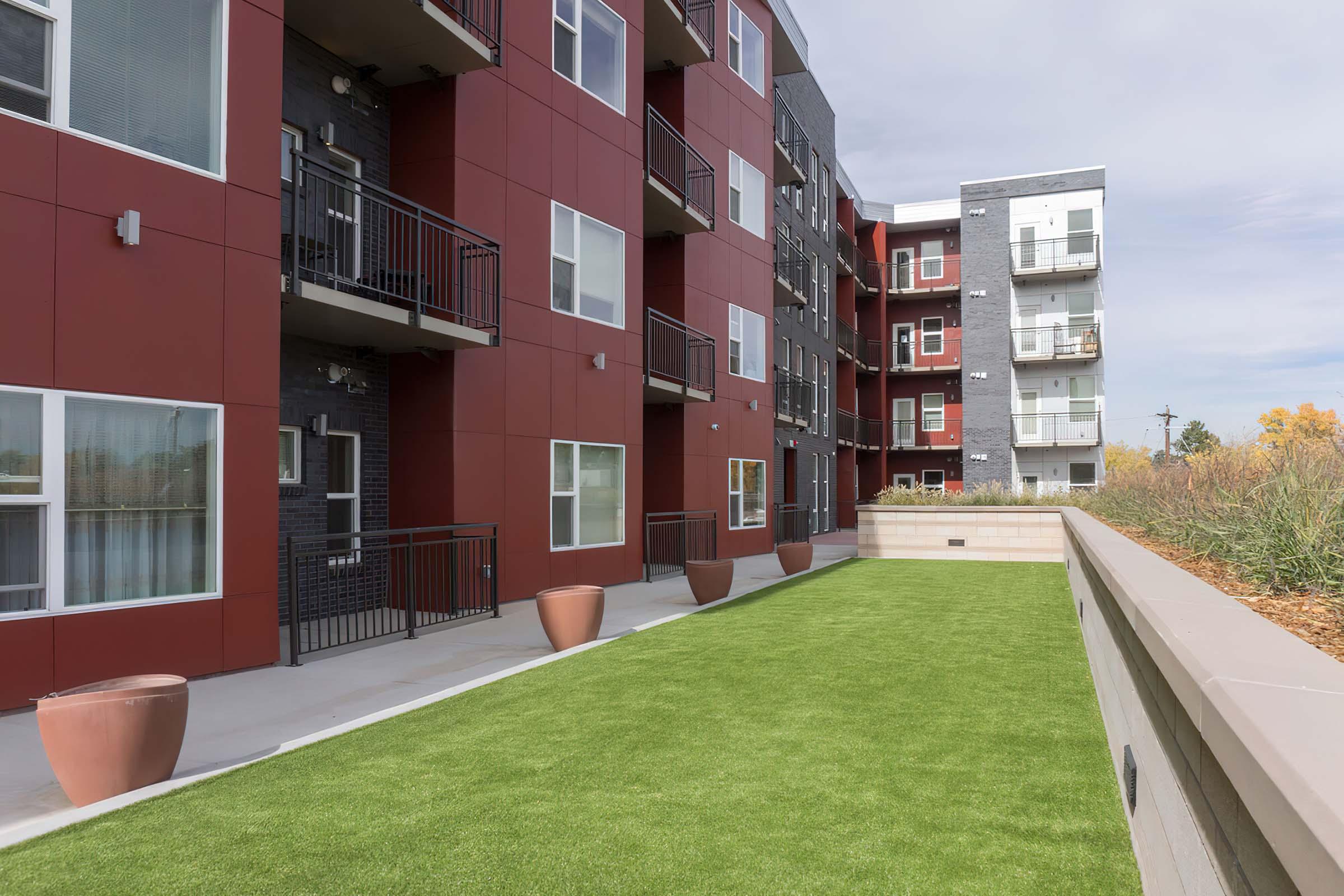 A modern apartment complex featuring red and gray building exteriors. There are balconies with railings and large windows. In the foreground, a well-maintained green lawn is visible, bordered by flower beds and flower pots, creating an inviting outdoor space. The sky is partly cloudy.