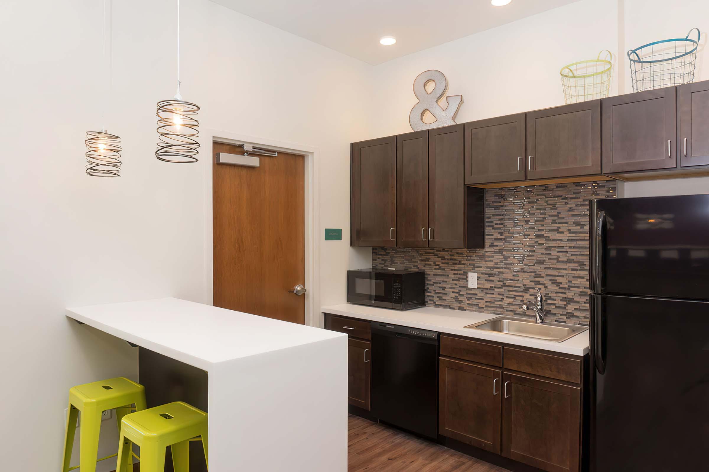 Modern kitchen with dark wood cabinets, a white countertop, and stainless steel appliances. Features a microwave, dishwasher, and a black refrigerator. Two bright green bar stools are positioned at a small breakfast bar. Stylish pendant lights hang from the ceiling, and decorative baskets add a pop of color to the space.