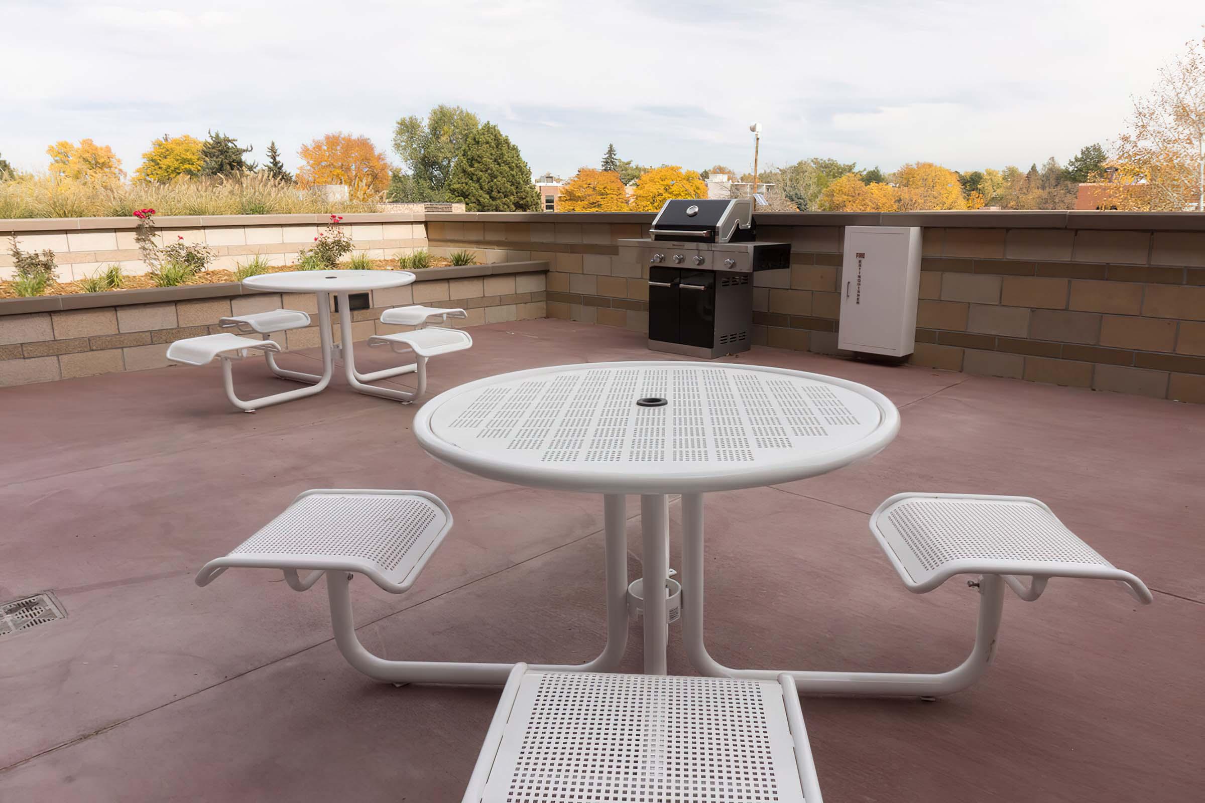 A rooftop patio featuring white metal tables and chairs, a barbecue grill, and a cooler. The area is surrounded by landscaped greenery with trees in autumn colors. The sky is partly cloudy, creating a pleasant outdoor setting.