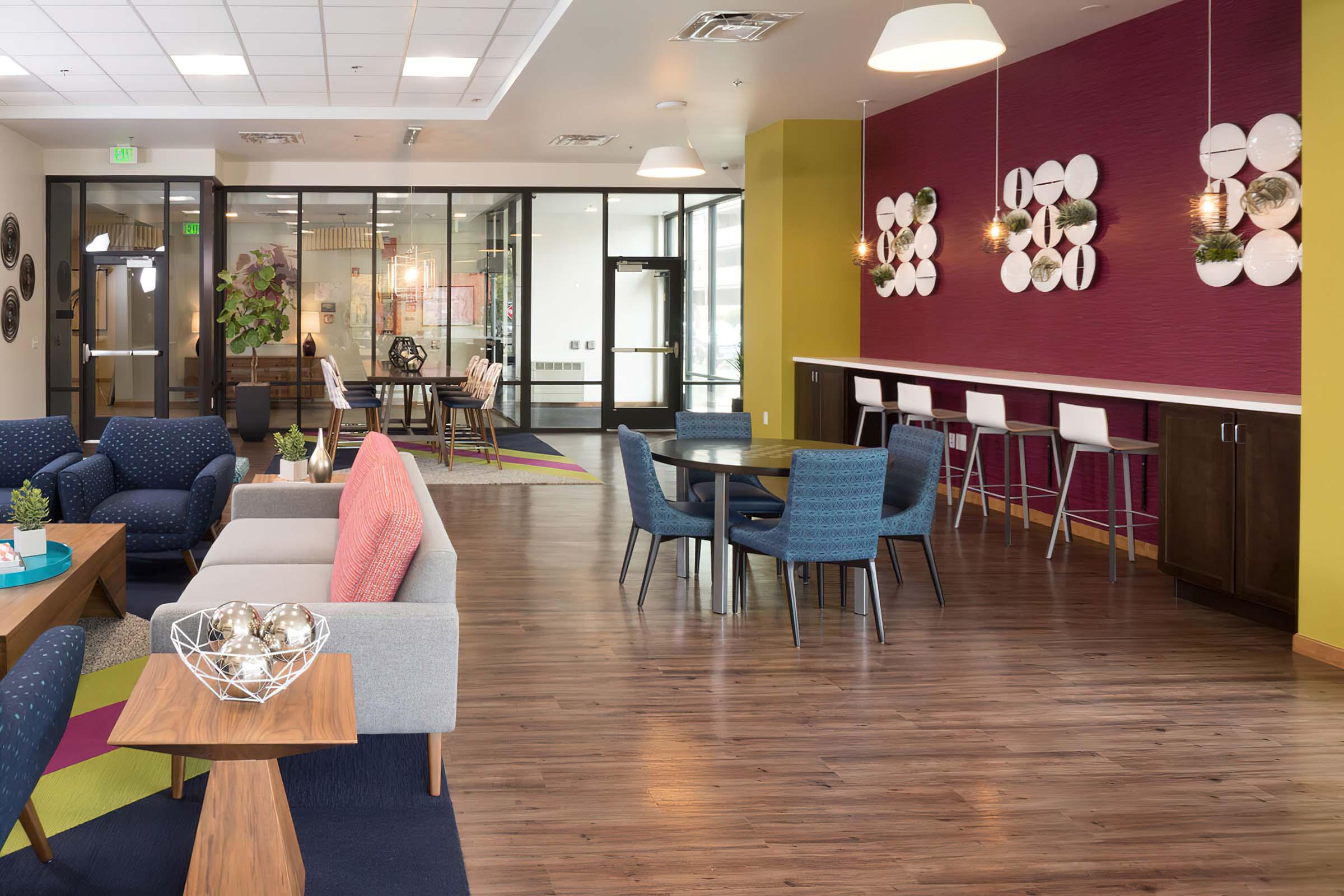 A modern, brightly lit lounge area featuring comfortable seating with colorful cushions, a dining table with chairs, and a bar counter. The walls are painted in vibrant colors, and decorative plates are displayed. Large windows provide natural light, creating an inviting atmosphere for socializing or relaxing.