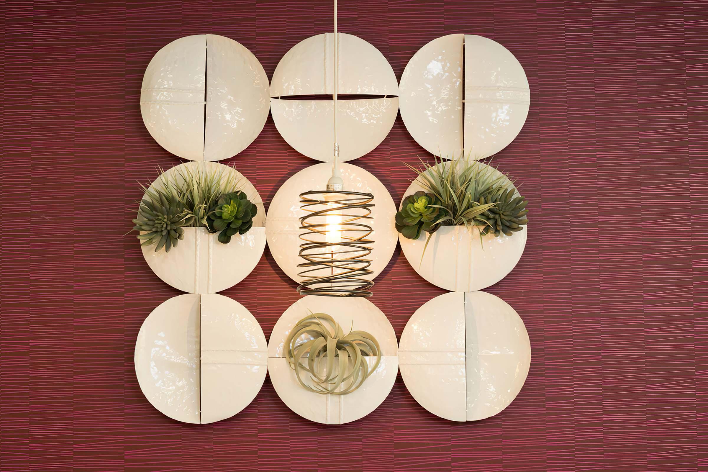 A modern wall art arrangement featuring circular white plates and small green plants, with a central light fixture consisting of interwoven metal spirals. The backdrop is a textured red wall, adding a vibrant contrast to the decor.