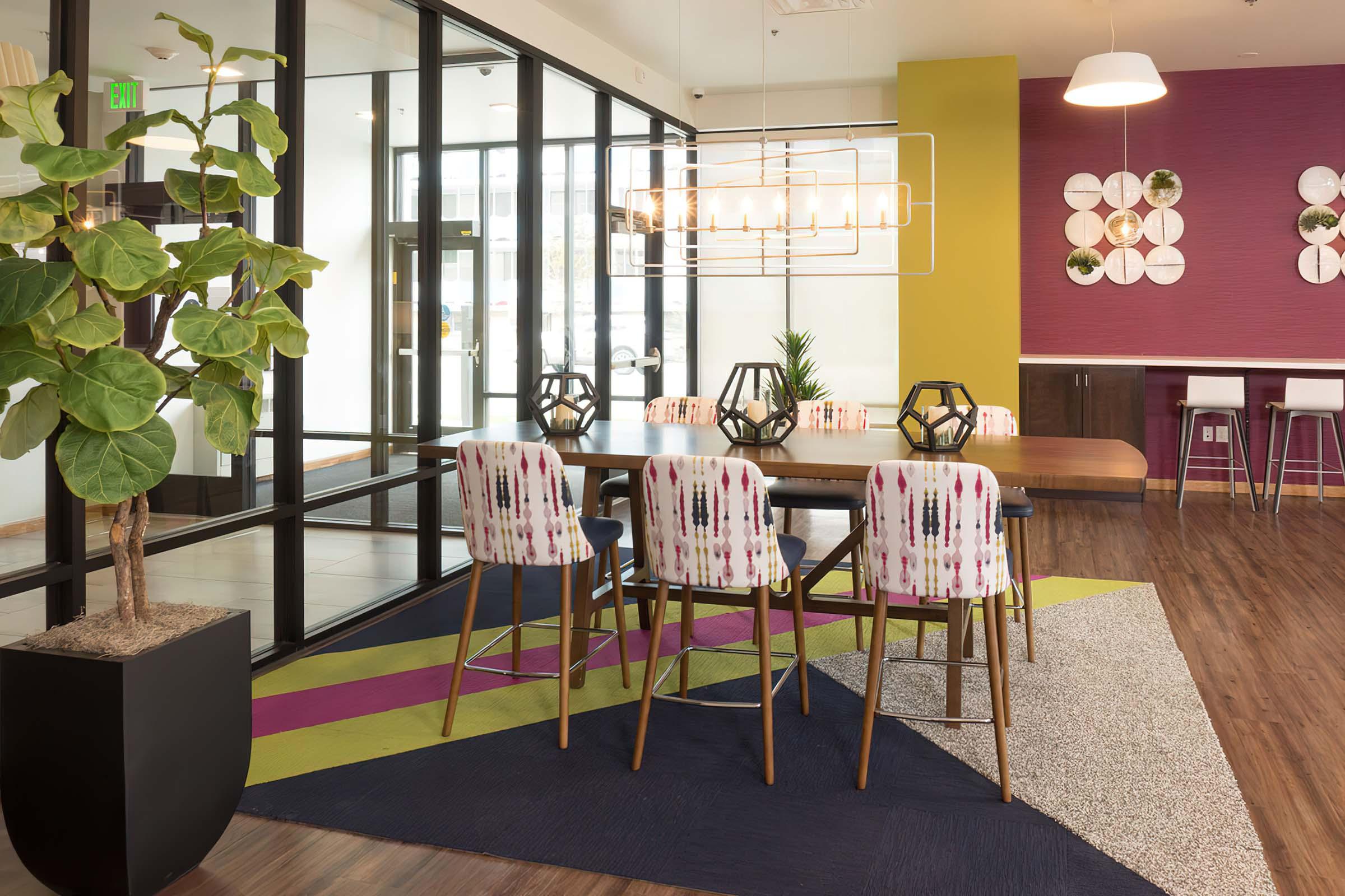 A modern dining area featuring a large wooden table surrounded by colorful chairs. The space includes a geometric light fixture overhead and is decorated with a variety of plants and wall art. Large windows provide natural light, while a vibrant area rug adds color to the floor.