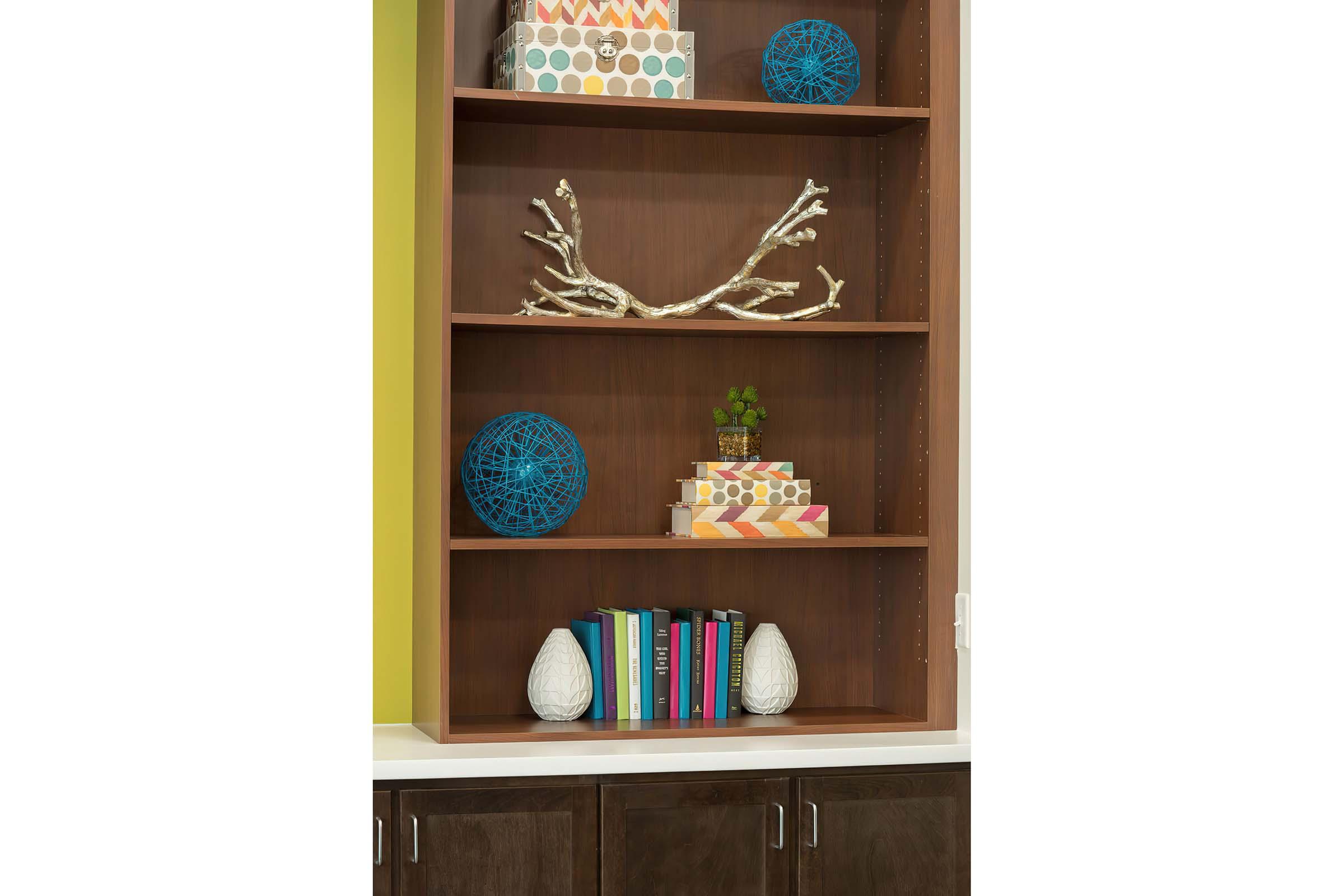 A wooden bookshelf with several shelves. Decor items include two blue decorative spheres, a small potted plant, a branch sculpture, colorful boxes, and a set of books. The background features a vibrant green wall, and the bottom part of the image shows a dark cabinet.