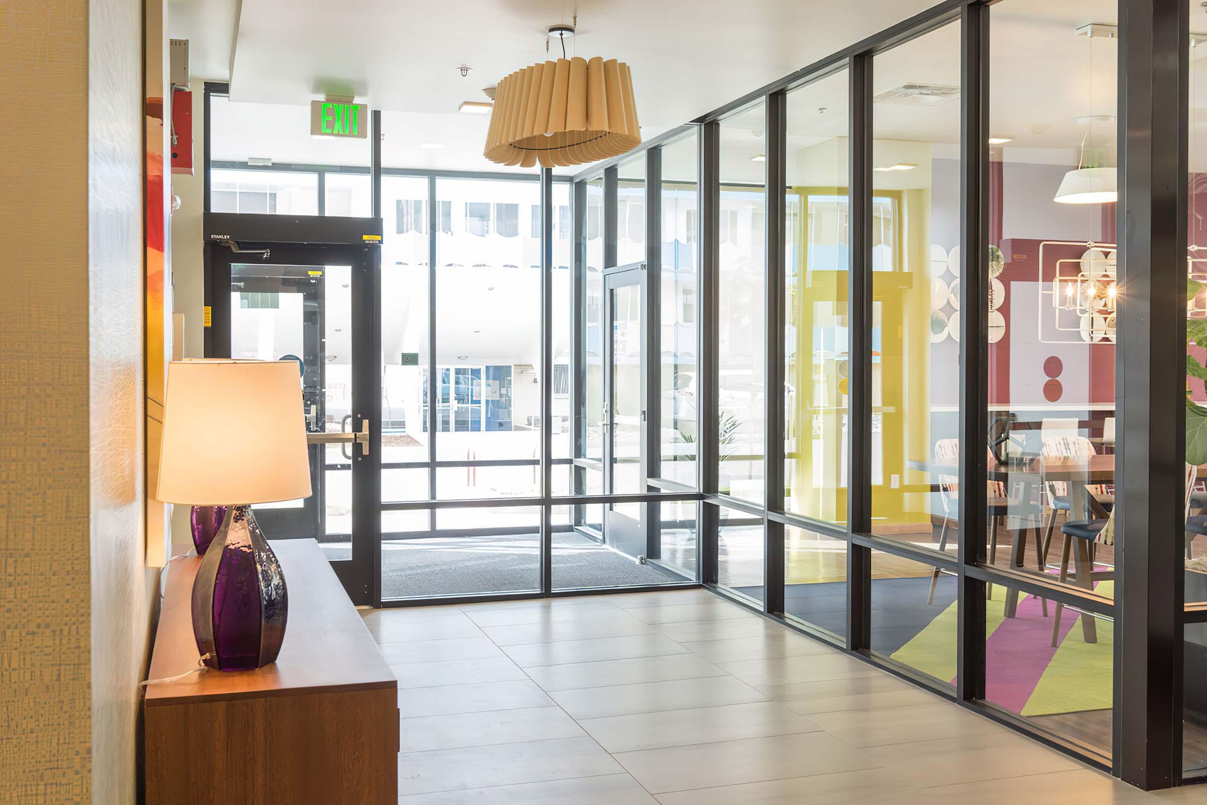 A modern lobby area featuring large glass windows, a decorative lamp on a wooden console table, and colorful accents on the walls. The entrance has double doors leading outside, while seating and dining areas can be seen in the background, enhancing the inviting atmosphere of the space.