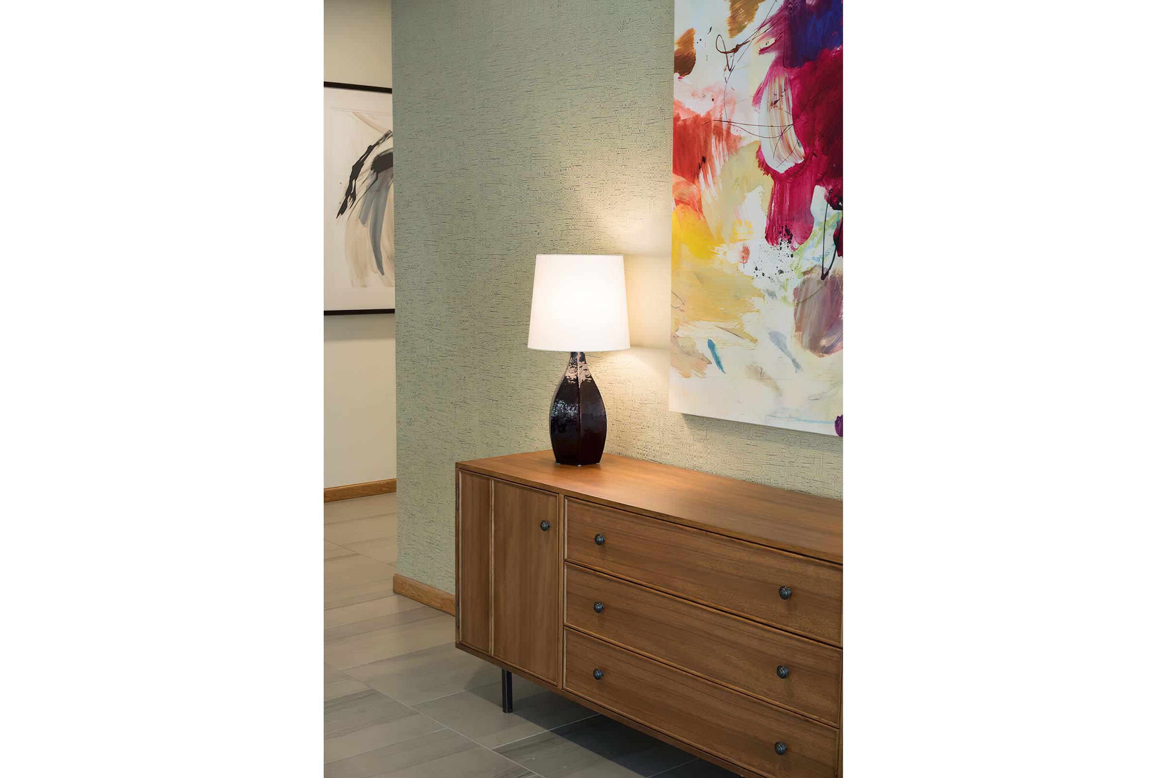 A modern interior scene featuring a wooden sideboard with three drawers and a dark vase-shaped lamp with a white lampshade. The wall behind has a textured green finish, while an abstract artwork with colorful splashes hangs nearby, adding a vibrant touch to the space.