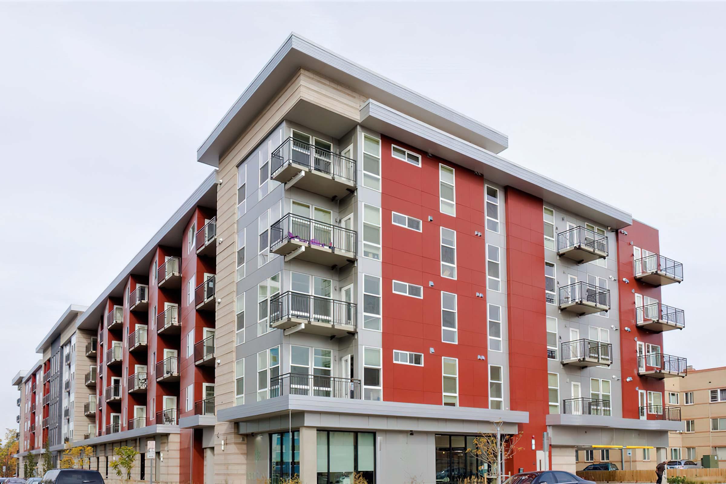 Modern apartment building with a combination of red and gray exterior. It features multiple balconies on each floor, large windows on the ground floor, and a contemporary architectural design. The surrounding area appears urban and well-maintained.