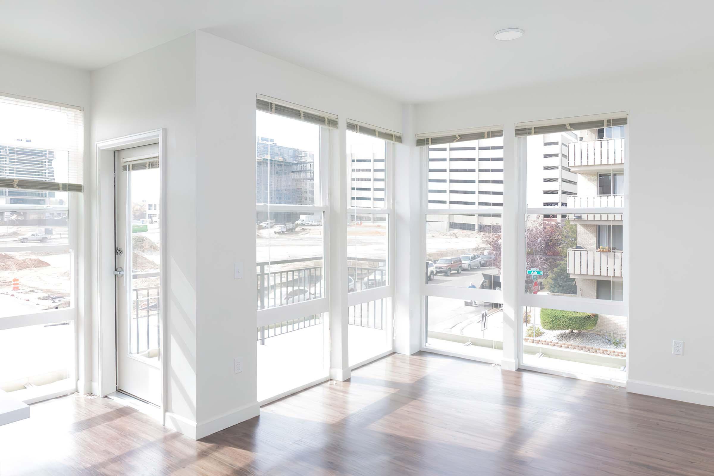 Bright, modern room with large windows allowing natural light, featuring light-colored walls and hardwood flooring. The view outside shows construction and nearby buildings. A door leads to an outdoor area. The space is uncluttered, emphasizing an open and airy feel.