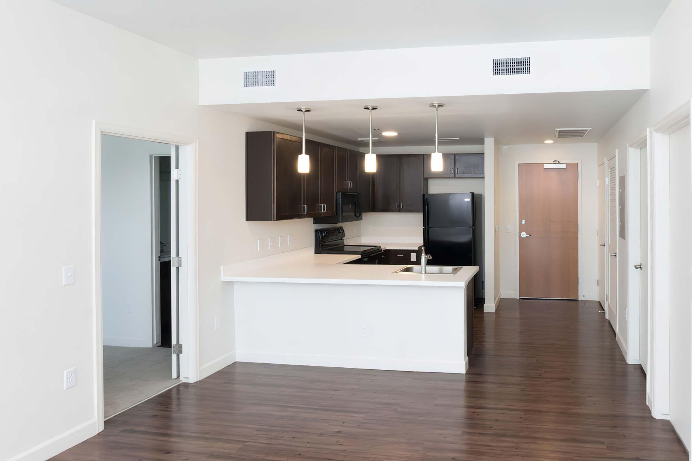 Modern kitchen area featuring dark wood cabinets, a white countertop, a black refrigerator, and pendant lighting. The space is open to a living area with wood flooring, and a doorway leads to another room. The walls are painted in neutral colors, creating a bright and inviting atmosphere.