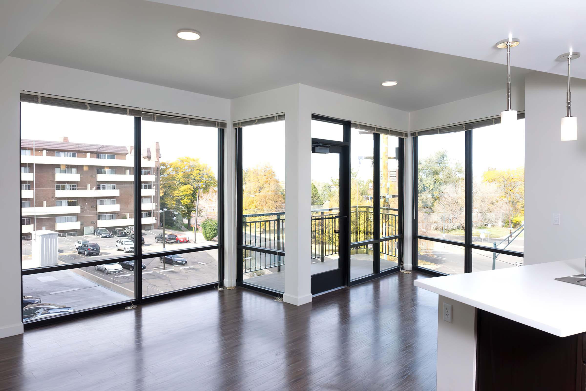 Bright and spacious living room featuring large windows that offer a view of nearby buildings and greenery. The modern design includes hardwood flooring and natural light flooding in, with a balcony door leading outside. Sleek light fixtures hang from the ceiling, enhancing the contemporary atmosphere.