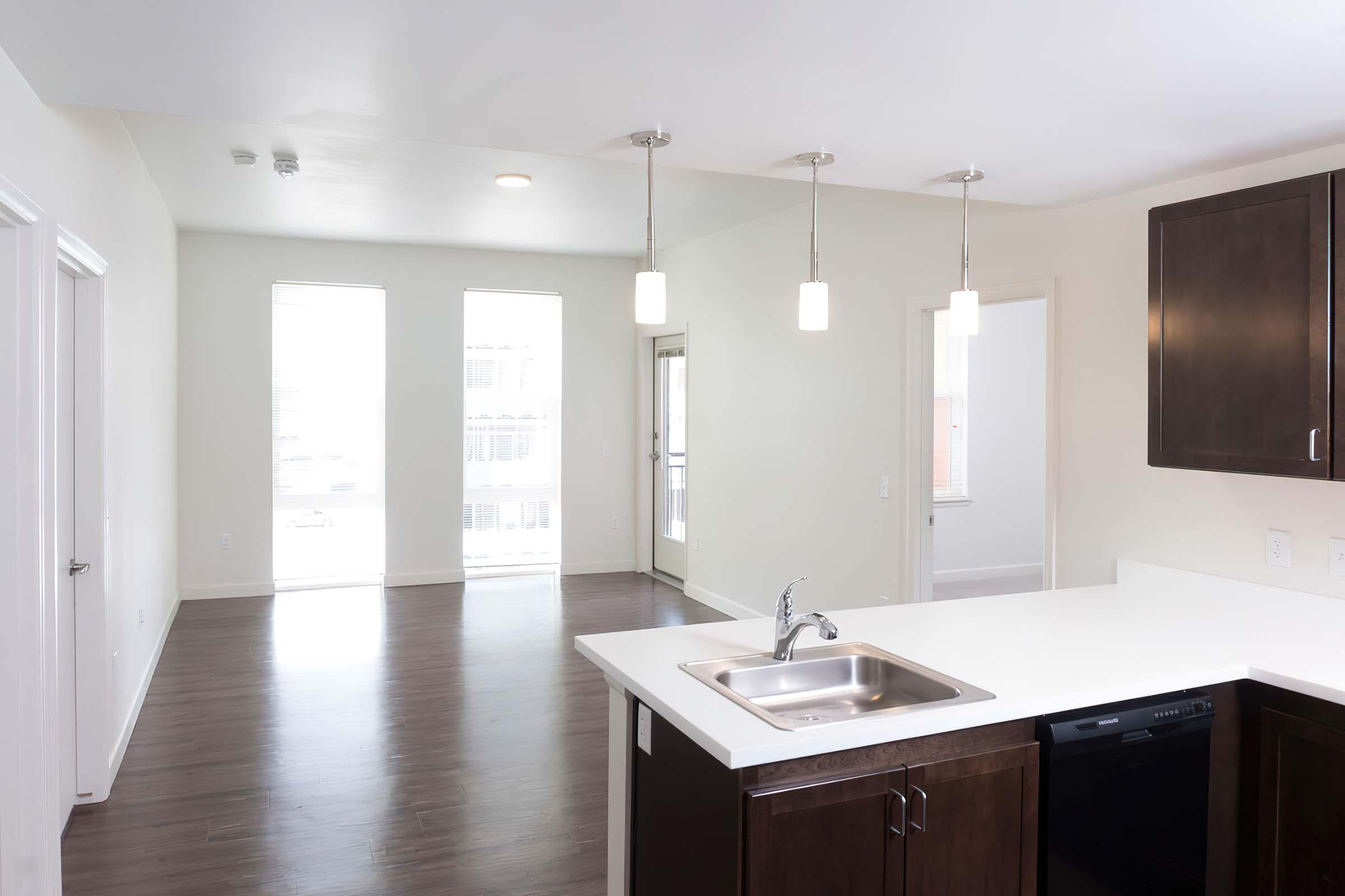 Modern kitchen with a white countertop, stainless steel sink, and dark wood cabinetry. Three pendant lights hang above the counter. Large windows in the background allow natural light into the open living space, which features hardwood flooring and light-colored walls.