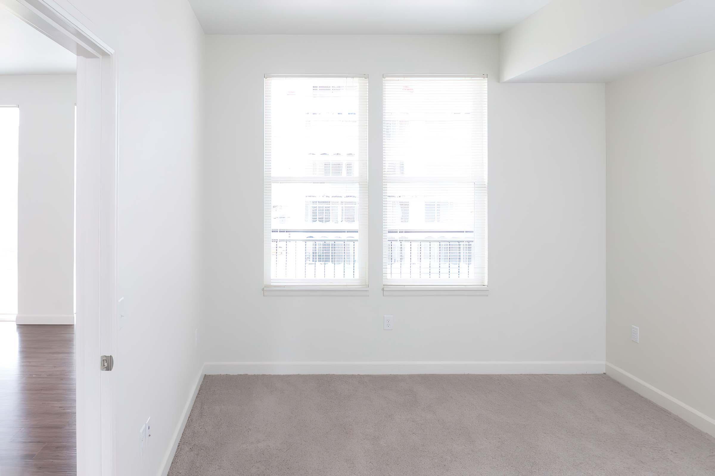 A bright, empty room featuring light-colored walls and a carpeted floor. Two large windows with blinds allow natural light to brighten the space. The room appears to have a doorway visible on the left. The overall atmosphere is spacious and inviting, ready for furniture and personal touches.