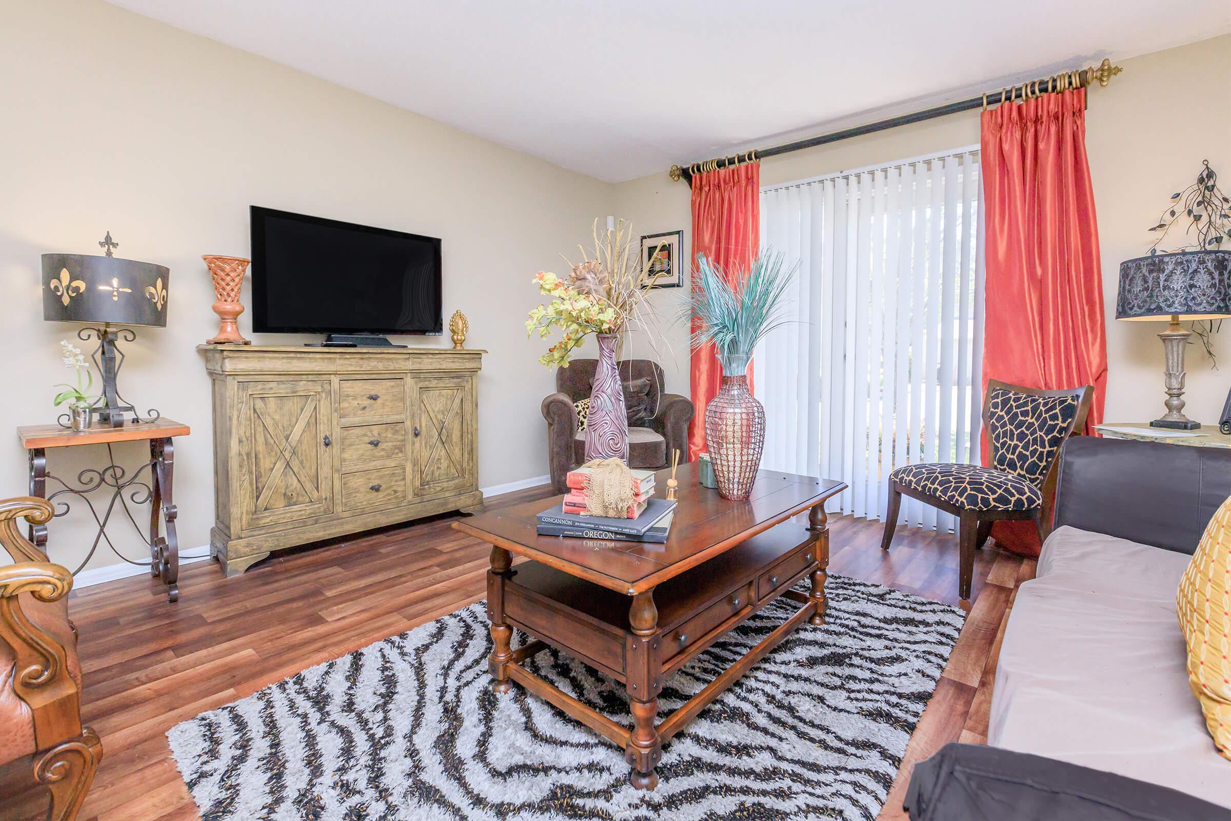 A cozy living room featuring a TV on a light-colored wall, a wooden console table, and decorative vases. A large area rug with a zebra pattern lies under a coffee table. The space is accented with orange and black curtains, a comfortable armchair, and stylish lamps, creating an inviting atmosphere.