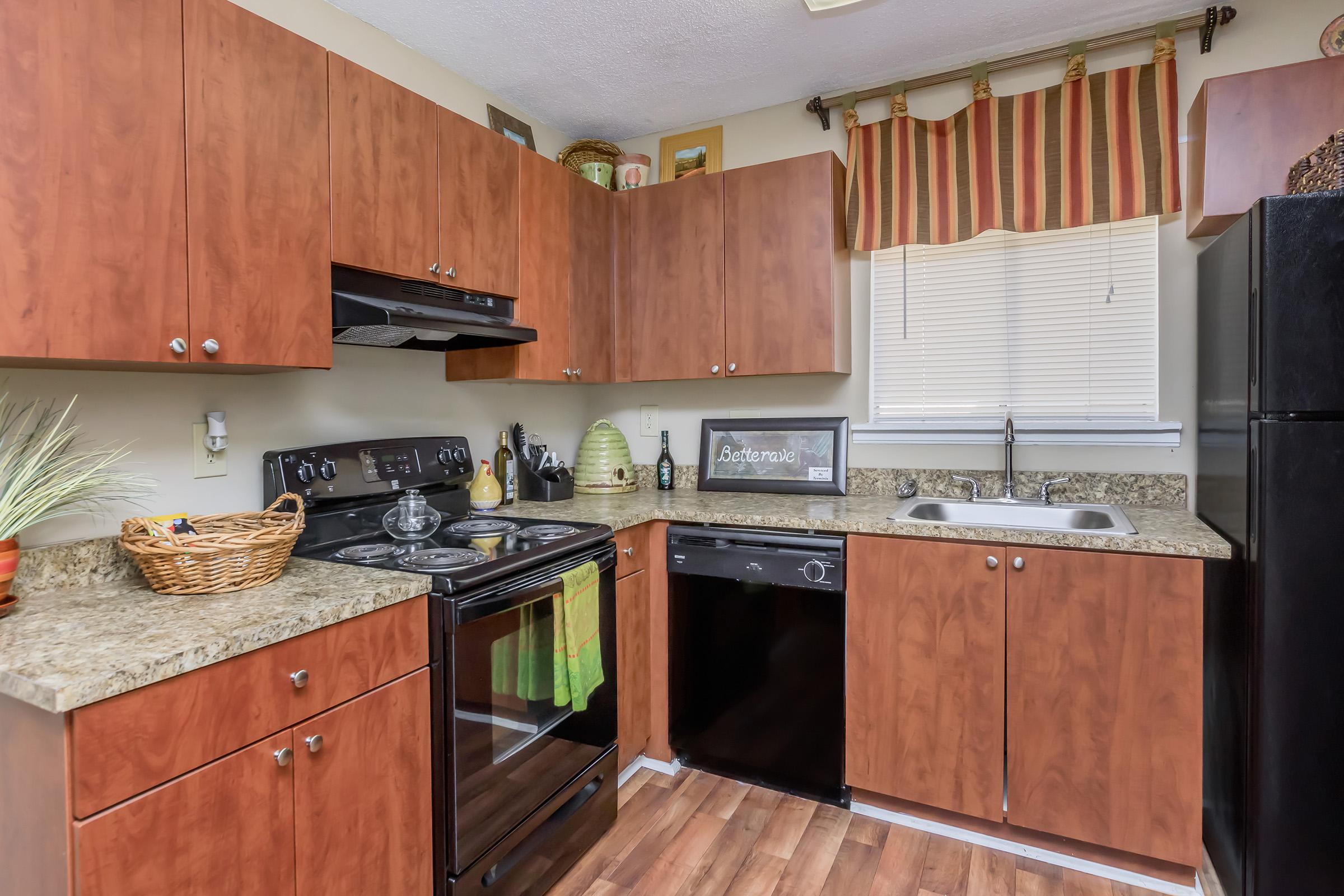 A cozy kitchen featuring wooden cabinetry, a black stove and dishwasher, and a granite countertop. There's a basket on the counter and decorative items on the shelves. Soft natural light streams in through a window with striped curtains, enhancing the warm atmosphere of the space.