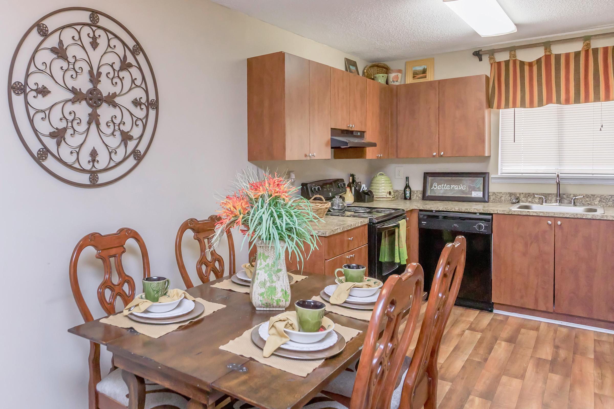 A cozy kitchen and dining area featuring a wooden table set for four with green and white dishes. A vase of colorful flowers sits at the center. The kitchen has brown cabinetry, a black stove, and a window with light curtains. Decorative wall art enhances the inviting atmosphere.