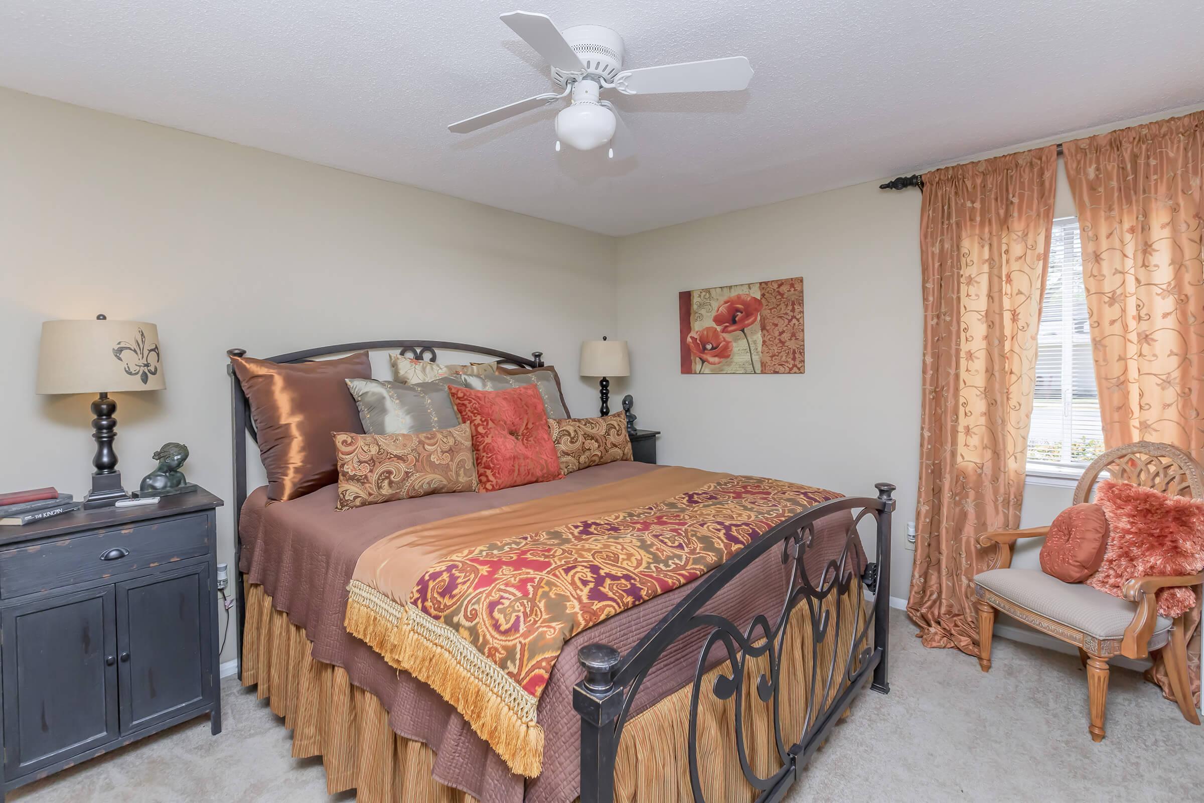 A cozy bedroom featuring a wrought iron bed with a richly adorned comforter and decorative pillows. There's a nightstand with lamps, an ornate window with orange curtains, and a small seating area with a wicker chair. A floral artwork adds a touch of elegance to the decor.