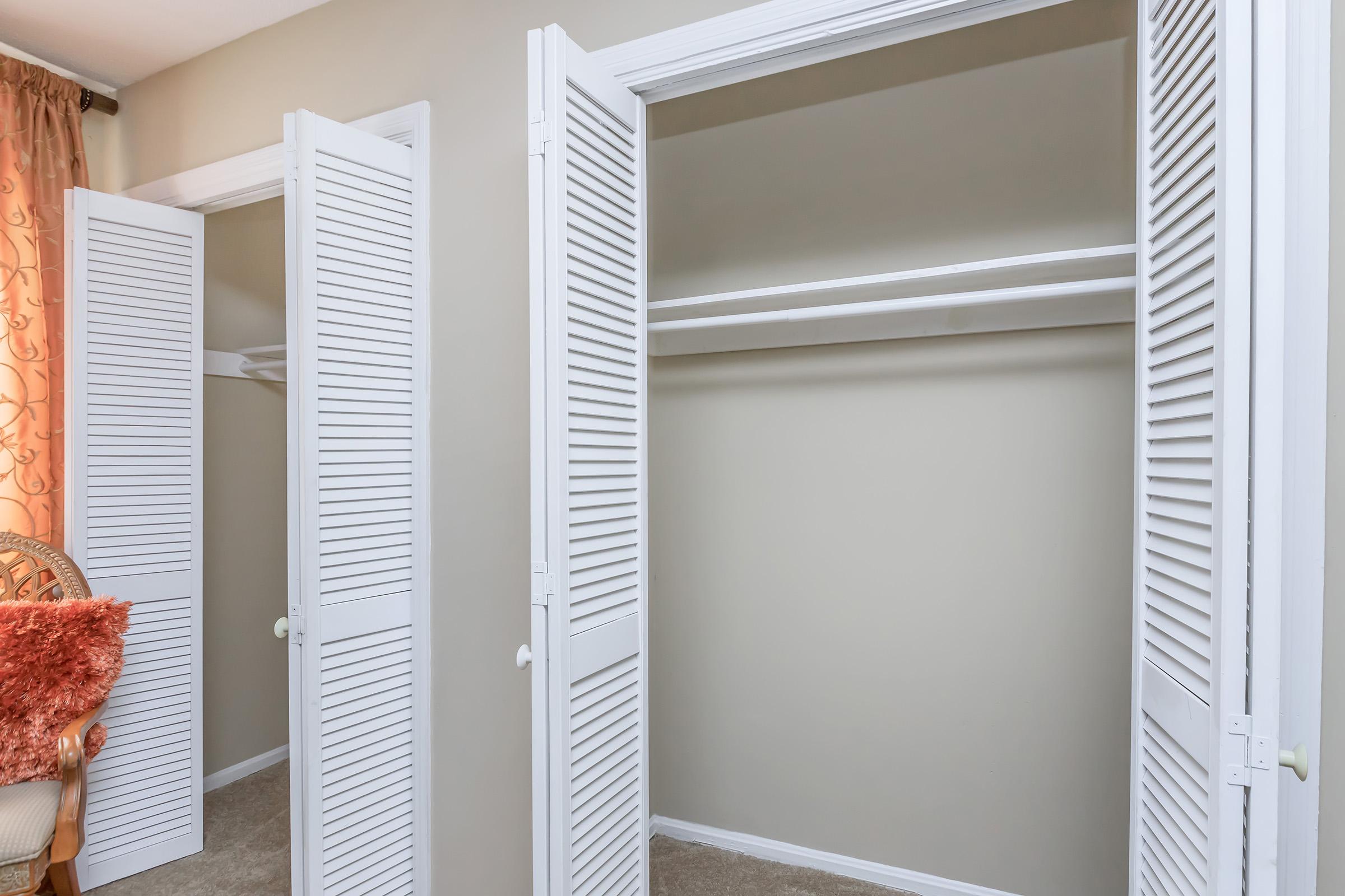 Two open closets with white louvered doors, showcasing the interior shelves against a neutral wall. The room has beige carpeting and a hint of a cozy chair with an orange cushion in the foreground, giving a warm atmosphere. Soft light filters in, enhancing the inviting feel of the space.