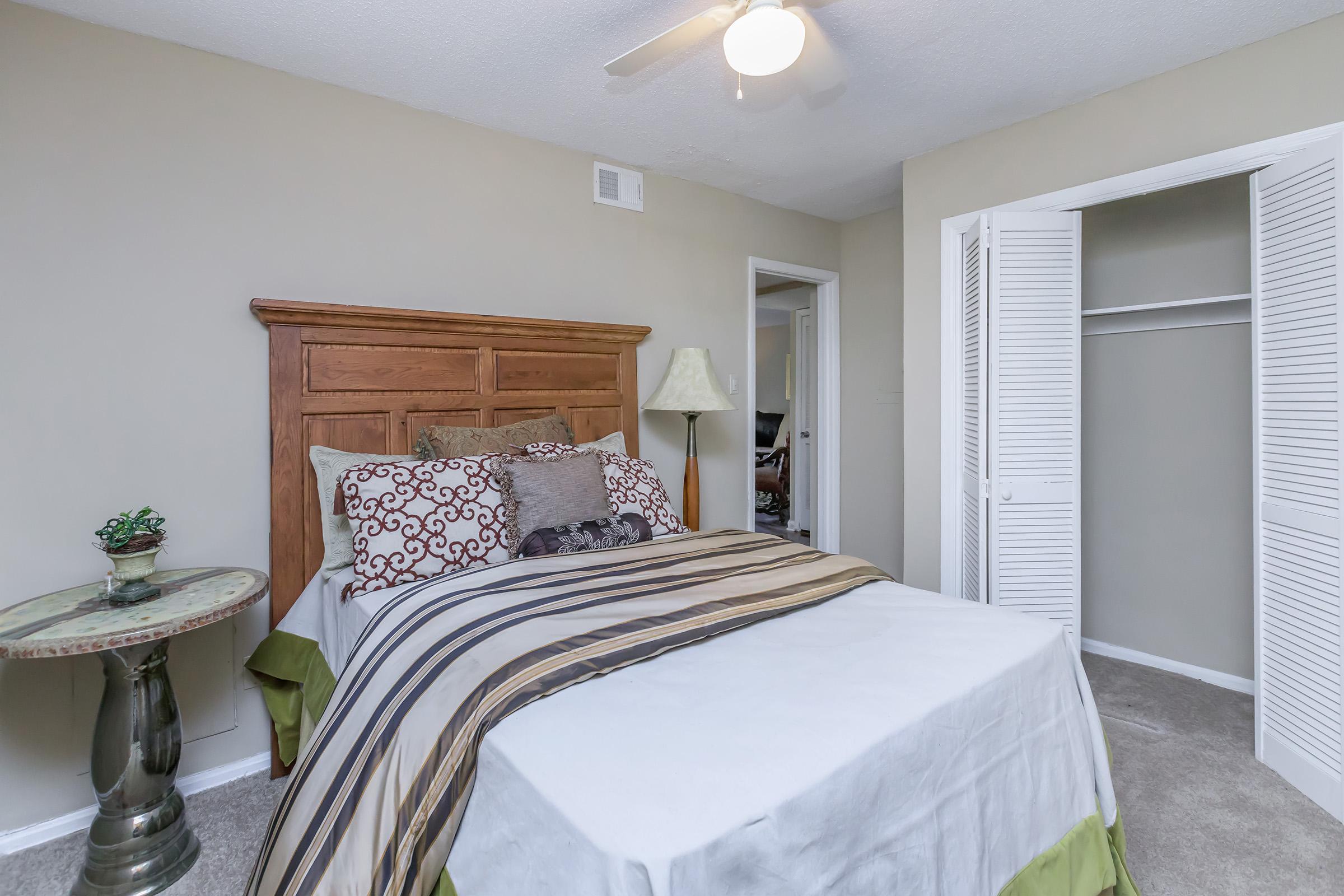 A cozy bedroom featuring a wooden bed frame with decorative pillows, a nightstand with a lamp, and a neutral color palette on the walls. A closet with sliding doors is visible, and the room is well-lit with natural light. The bedding includes a patterned throw and a green accent on the edge.