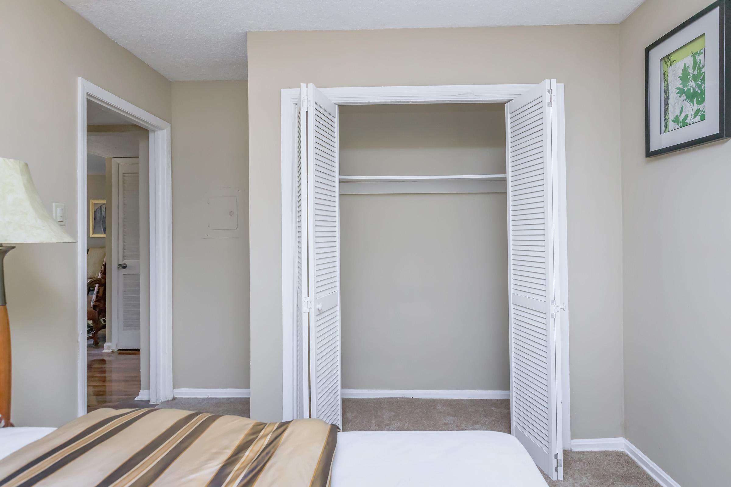 A well-lit bedroom featuring a neatly made bed with striped bedding. On the left, a lamp and a partially visible wall. A closet with louvered doors stands open, revealing an empty space inside. The walls are painted a neutral color, and there is a framed picture hanging near the closet.