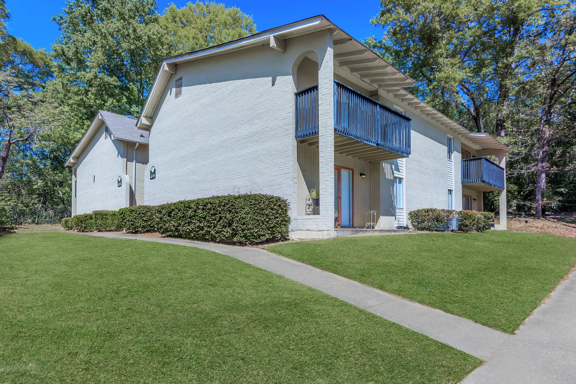 Two-story residential building with a light-colored exterior, featuring blue railings on the balconies. The property is surrounded by neatly trimmed green grass and bushes, with a concrete pathway leading up to the entrance. Trees provide a natural backdrop in a sunny environment.