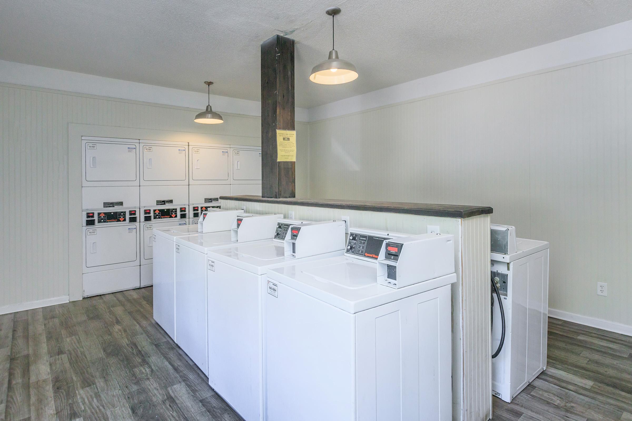 A clean, well-lit laundry room featuring several white washing machines and dryers lined up against a wall, with overhead pendant lights and a neutral-toned interior. A row of lockers or storage units is visible in the background, providing additional utility.