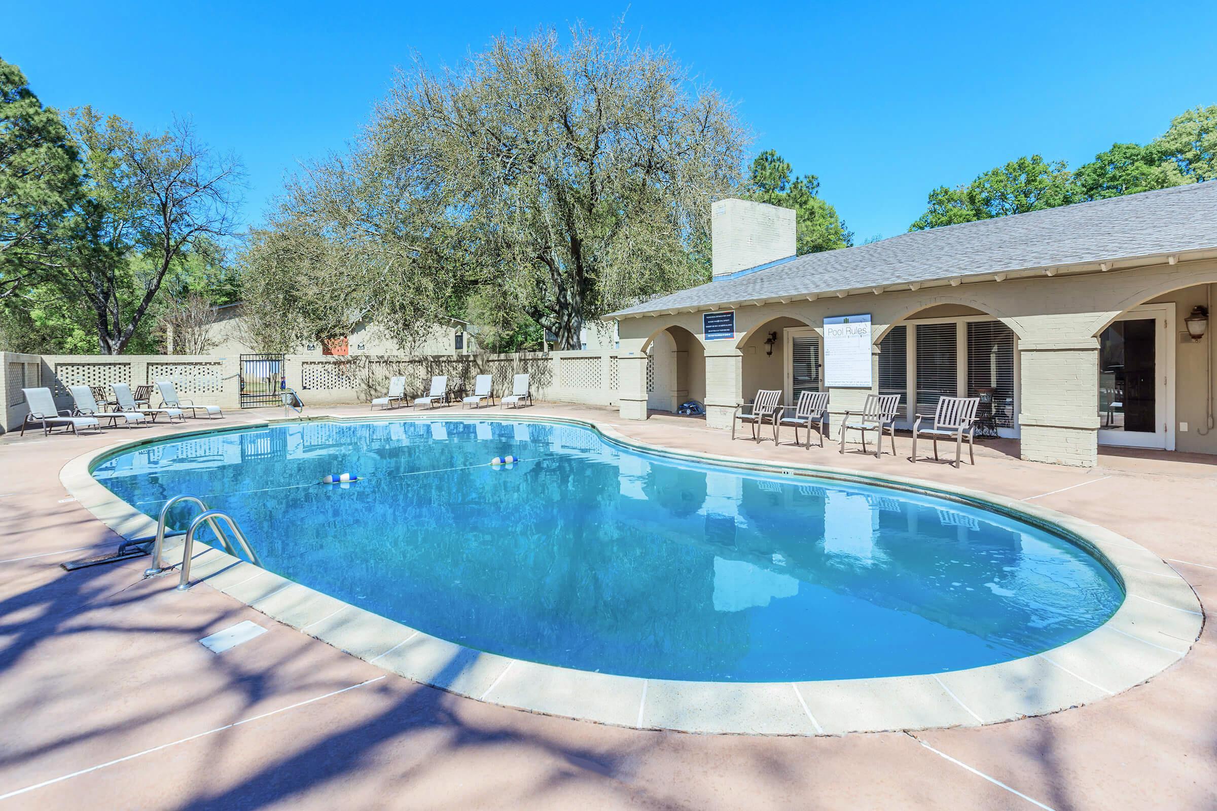 A clear blue swimming pool surrounded by a concrete deck, with several lounge chairs positioned nearby. Tall trees provide shade in the background, and a clubhouse or building is visible to the right. The sky is bright and sunny, creating a vibrant outdoor atmosphere.