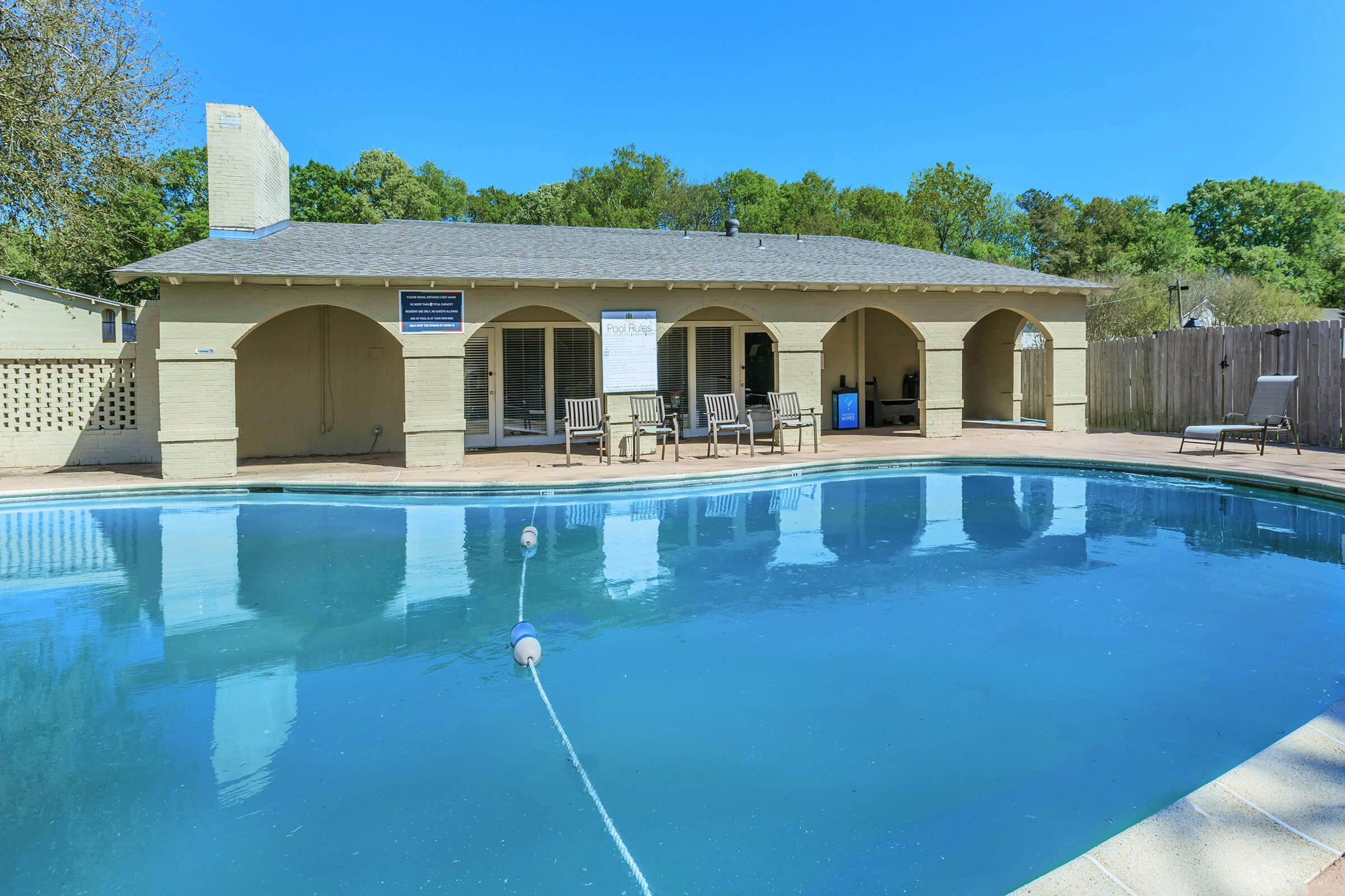 A serene view of a swimming pool with clear blue water, surrounded by a beige building featuring arched entrances. The building has a sign and several outdoor chairs positioned on a patio area. Lush greenery is visible in the background, and the sky is clear and bright.