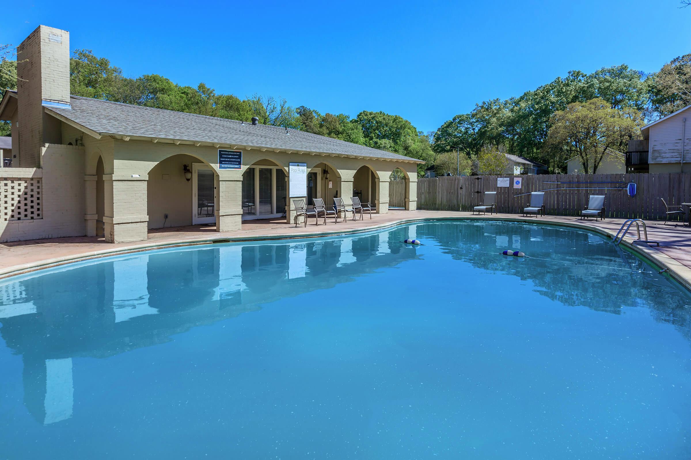 A clear, inviting swimming pool surrounded by lounge chairs and a sunlit patio. In the background, there are trees and a building with an outdoor area. The sky is bright blue, enhancing the tranquil atmosphere of the setting.