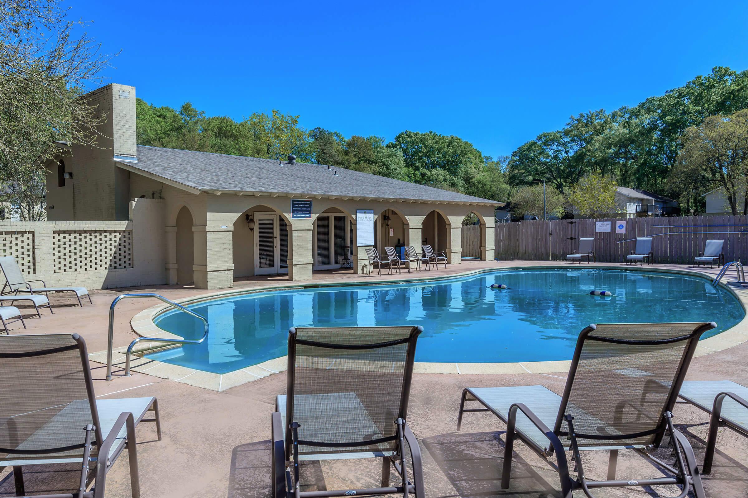 A serene outdoor pool area featuring a clear blue pool surrounded by lounge chairs. In the background, there is a beige building with arched architecture. Lush green trees line the perimeter, and the sky is bright blue, creating a tranquil atmosphere ideal for relaxation.
