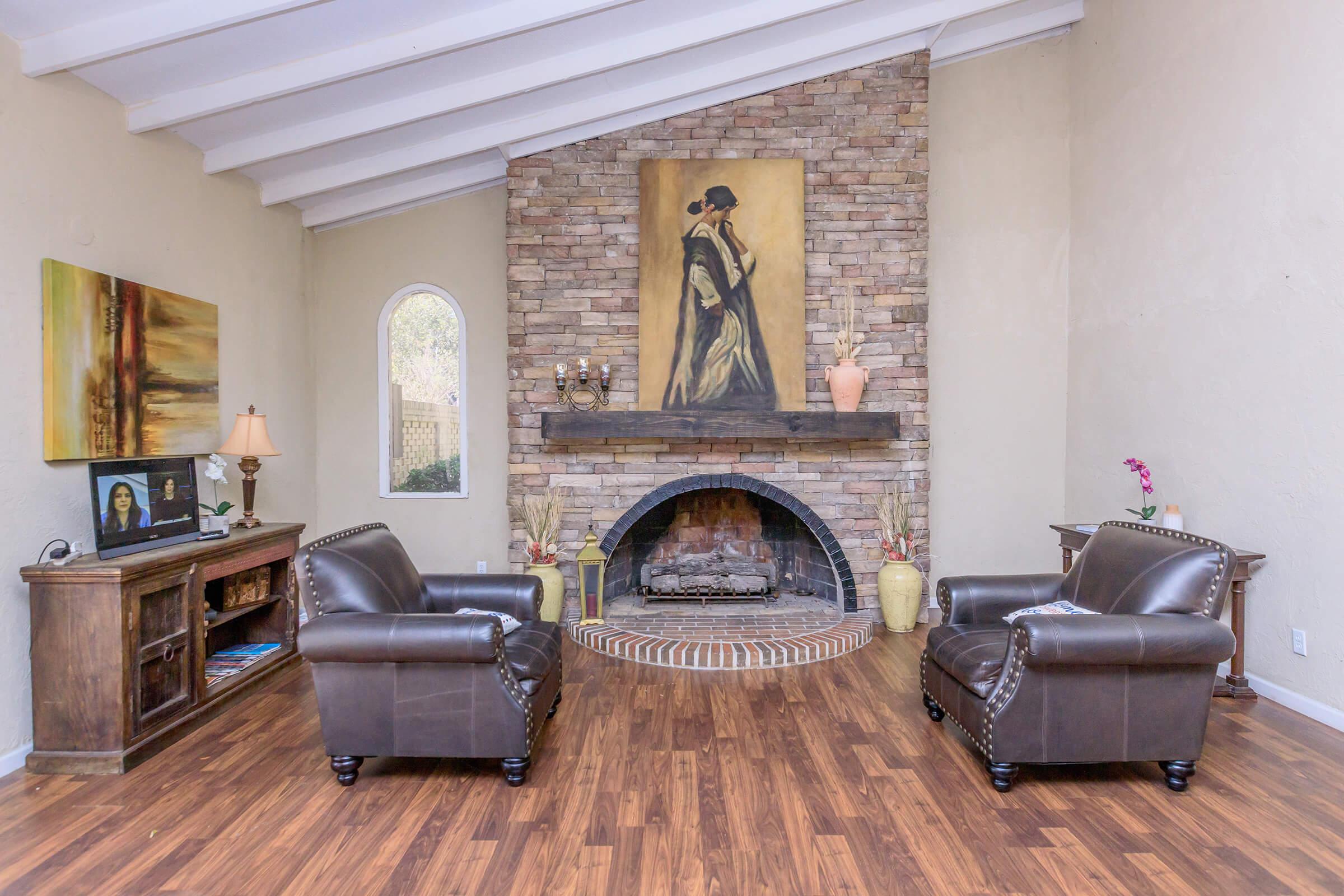A cozy living room featuring two brown leather armchairs facing a traditional stone fireplace. Above the fireplace is a large painting of a woman. A wooden TV stand with a lamp and decorative items is on one side, while potted plants add a touch of color to the warm, inviting space with wooden flooring.