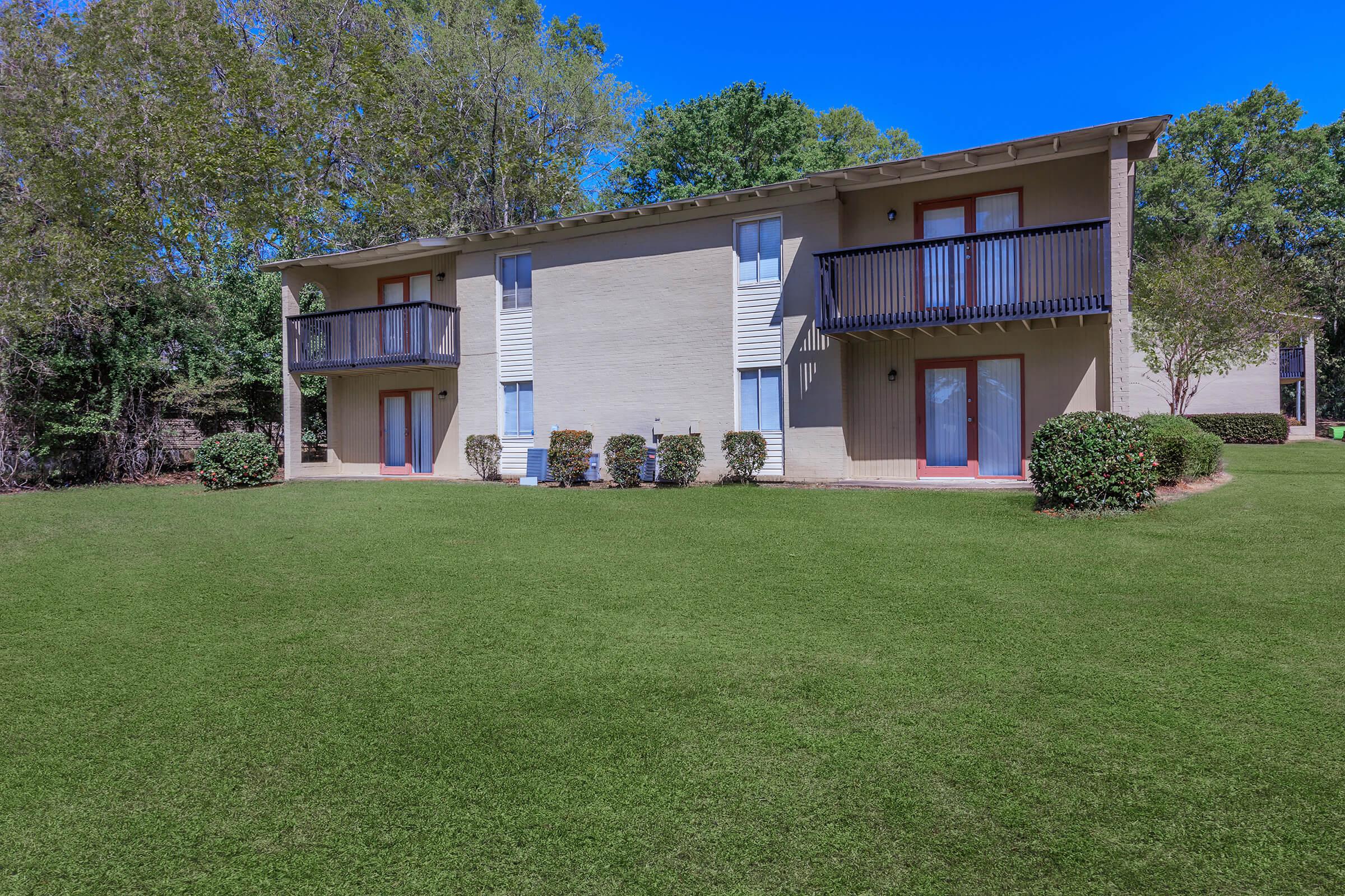 A two-story apartment building with a light-colored exterior and blue accents. The building features balconies on both upper levels, surrounded by well-maintained green grass and shrubbery. The background includes trees under a clear blue sky.