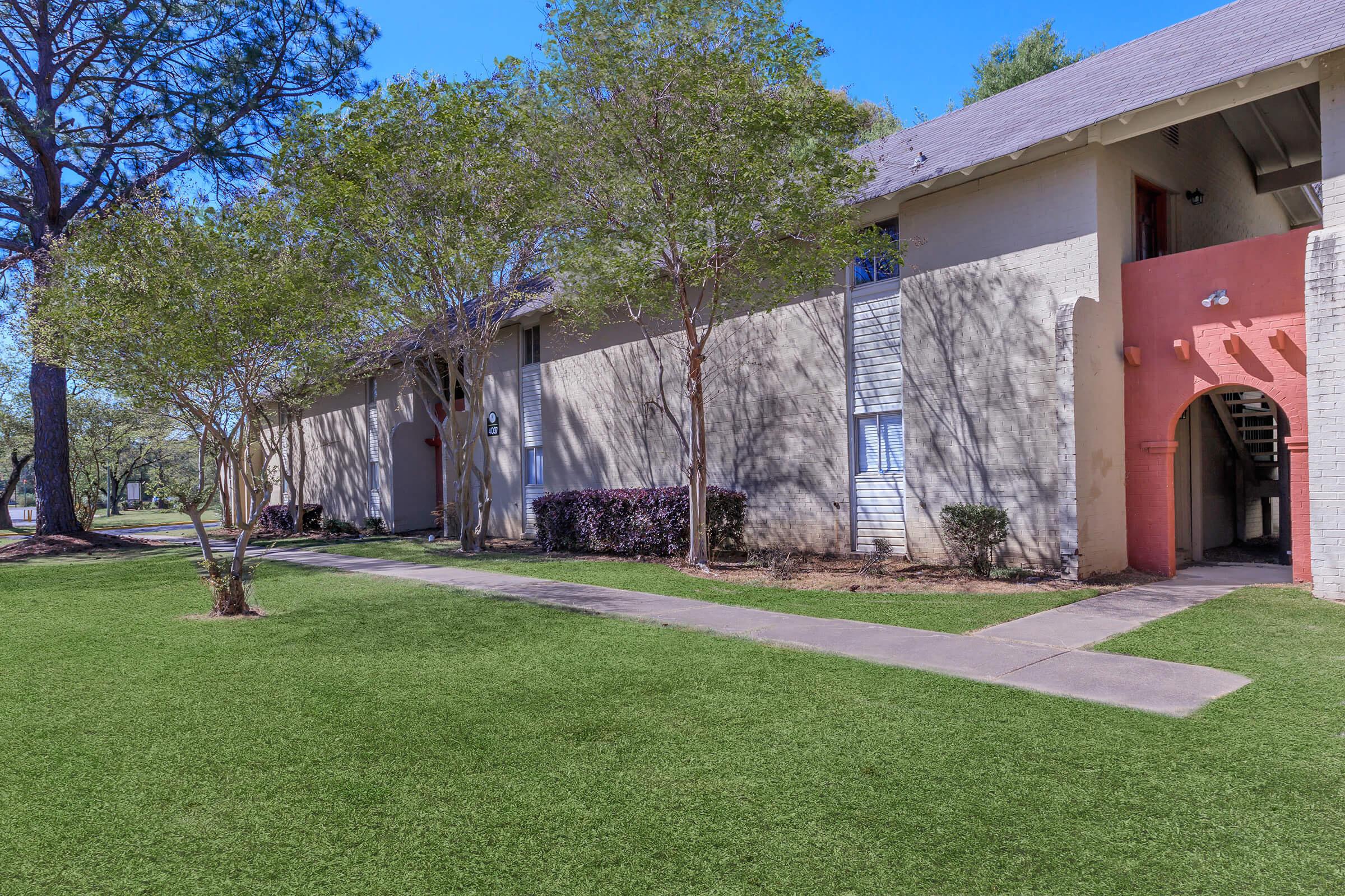 A view of a residential building surrounded by grass and trees. The building features a mix of light-colored walls with a pink accent, large windows, and an entrance with stairs. The landscape is well-maintained, providing a pleasant outdoor environment.