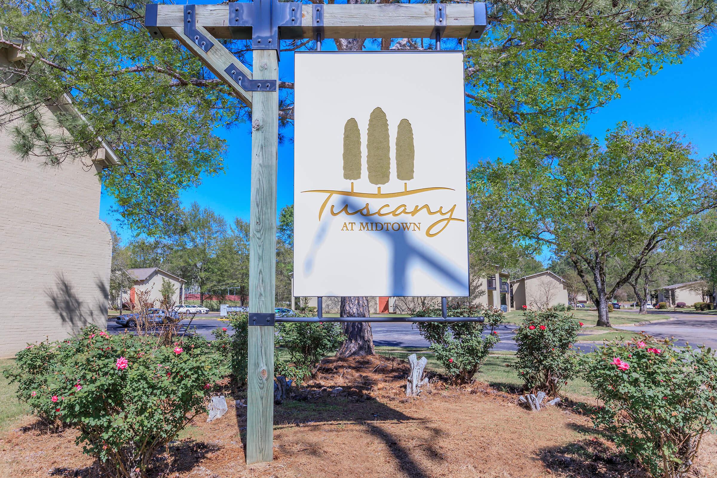 A wooden signpost featuring a white banner with the text "Tuscany at Midtown" in elegant font, accompanied by three green tree silhouettes. The background includes manicured shrubs with pink flowers and a clear blue sky, suggesting a serene residential area.