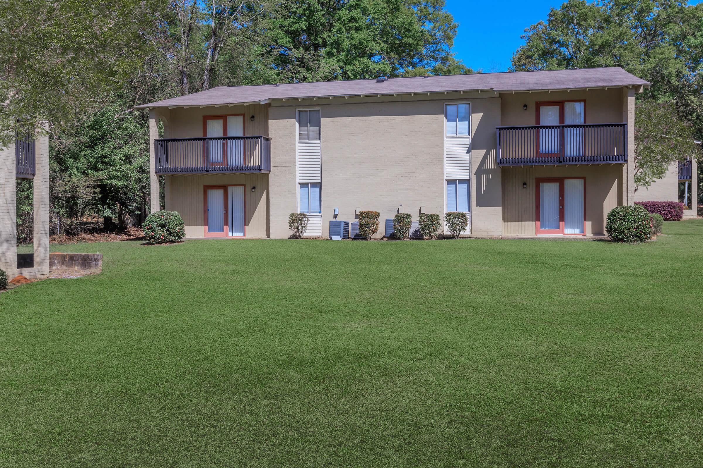 Two-story apartment building with a beige exterior, featuring two balconies on the upper level. The building is surrounded by a neatly maintained green lawn and small bushes. Trees in the background indicate a suburban setting under a clear blue sky.