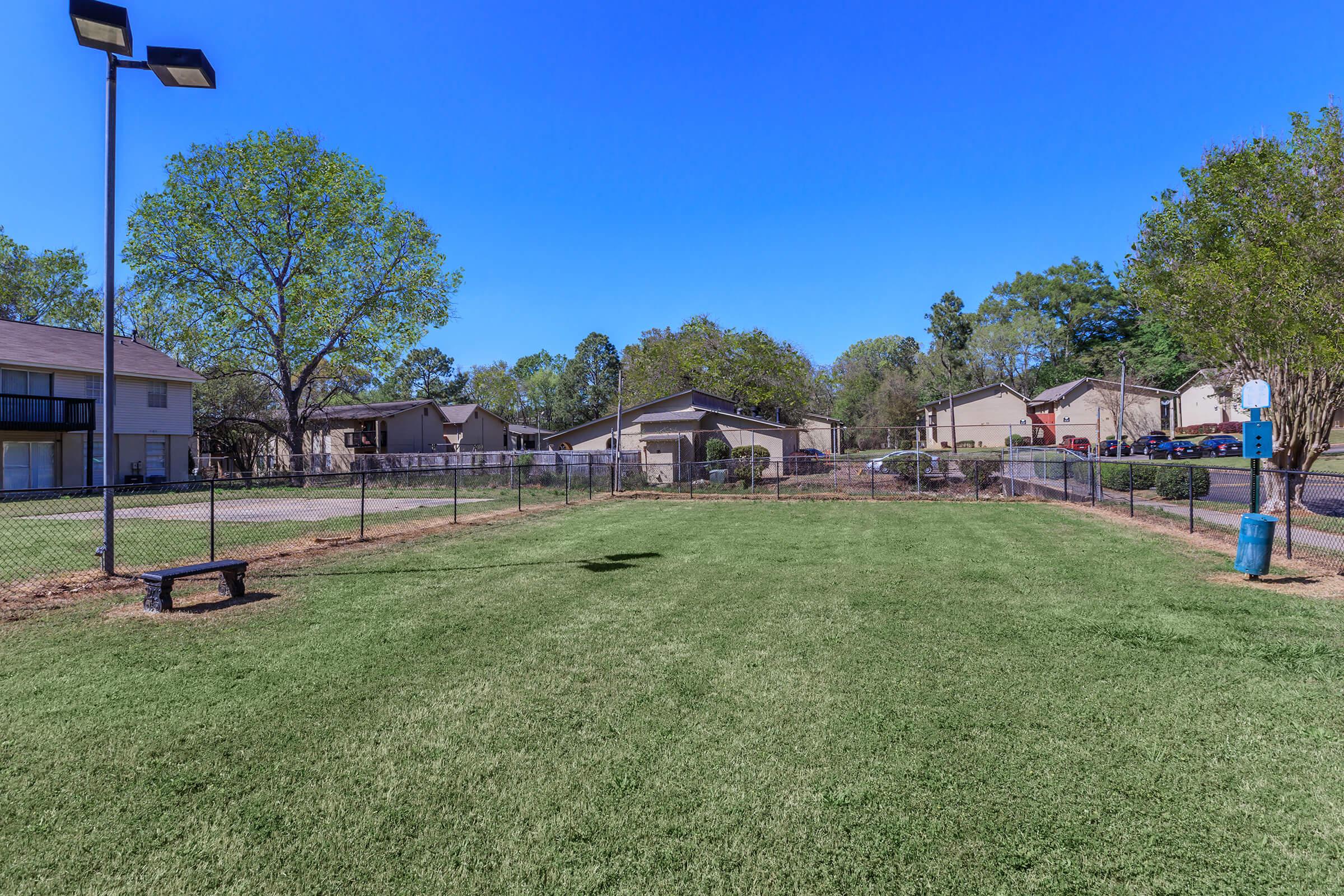 A bright, sunny day at an open green space enclosed by a chain-link fence. The area features well-maintained grass, benches, and trees in the background. Nearby residential buildings and parking areas are visible, contributing to a community atmosphere. Ideal for outdoor activities and relaxation.