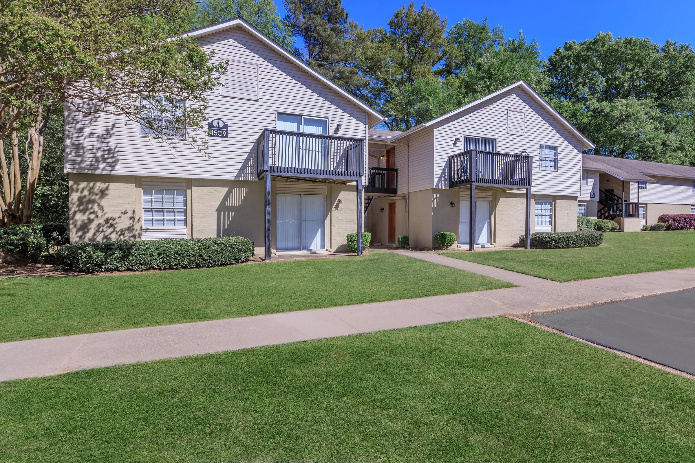 A serene view of a residential apartment community featuring two two-story buildings with balconies, surrounded by neatly trimmed lawns and shrubs. The sky is clear and blue, adding to the peaceful atmosphere of the neighborhood. A paved walkway runs alongside the buildings, leading to the entrances.
