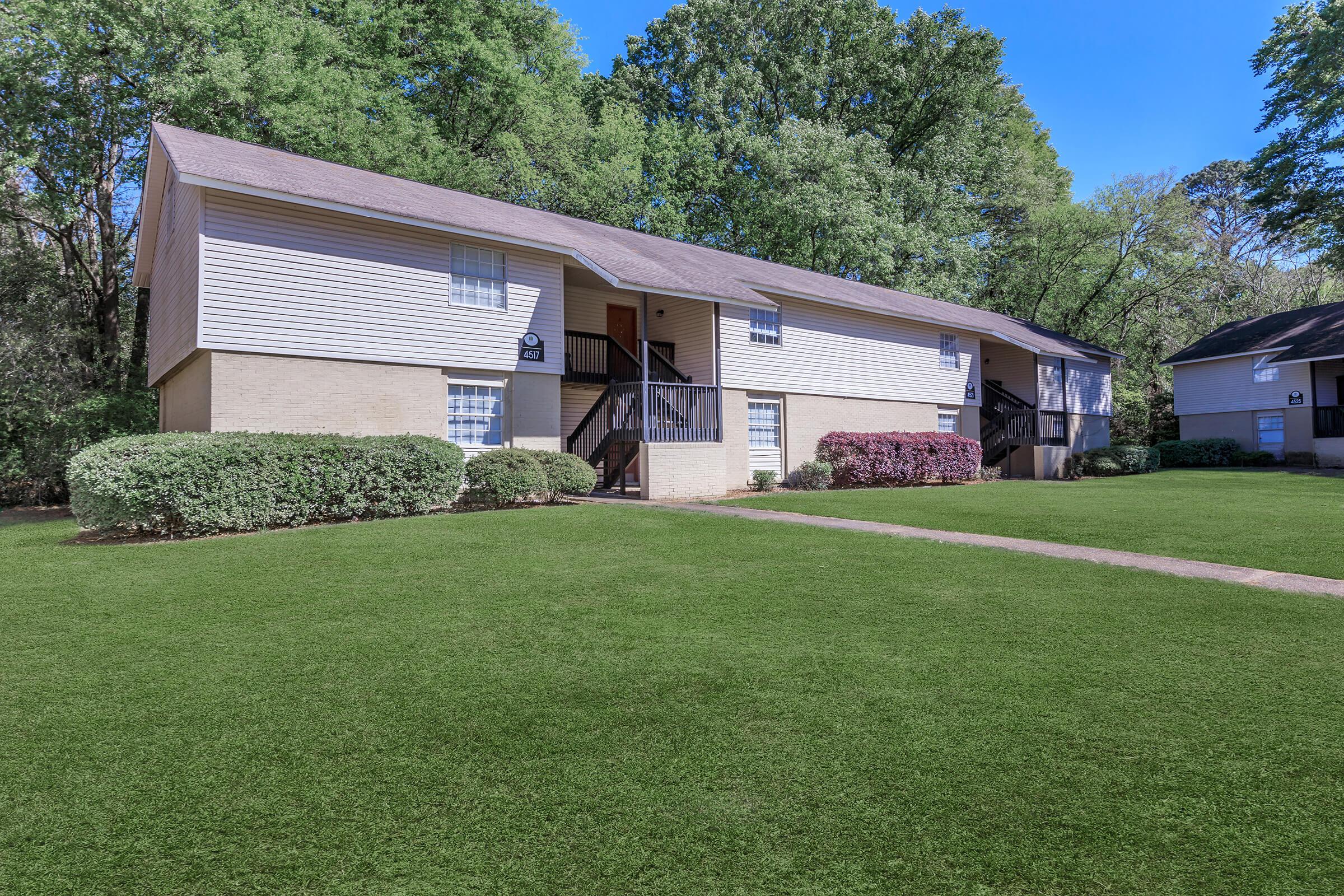 Two-story residential apartment building surrounded by well-maintained green lawns and shrubbery. The building features a light-colored exterior with a dark staircase and multiple windows. Trees in the background provide a serene, natural setting. Clear blue sky adds to the inviting atmosphere.