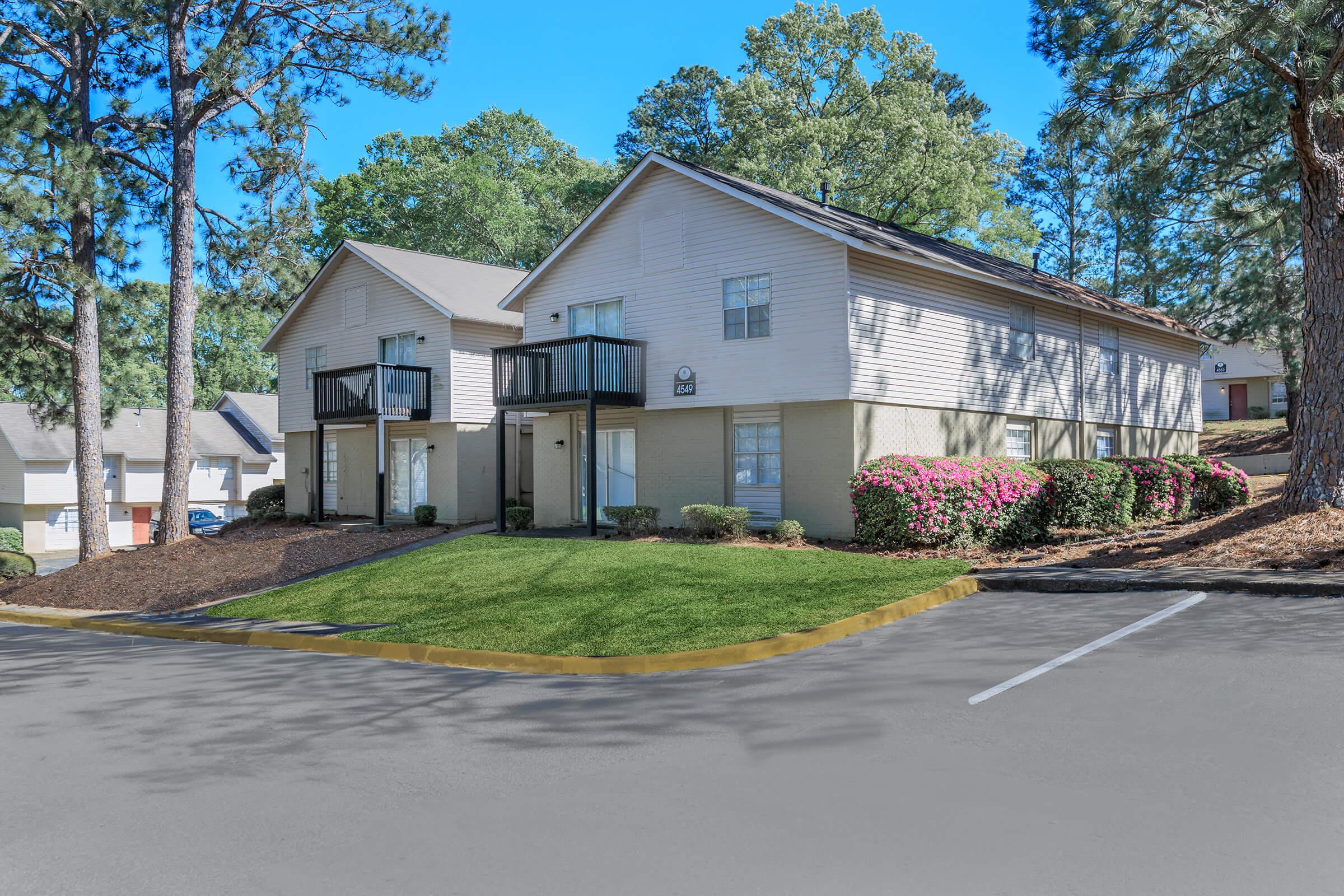 A well-maintained two-story apartment building surrounded by lush trees and colorful flower beds. The landscape features a manicured lawn, a paved parking area, and balconies on the upper level. Bright blue sky complements the serene environment, creating an inviting atmosphere for residents.