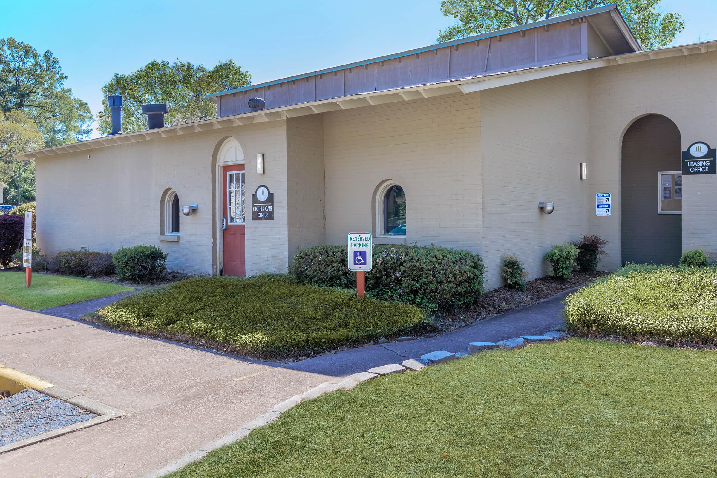 A modern building with a light-colored exterior and a red door. There is a sign for a leasing office and accessible parking. The area features neatly trimmed bushes and a landscaped yard with grass and stones along the walkway, set against a backdrop of trees.