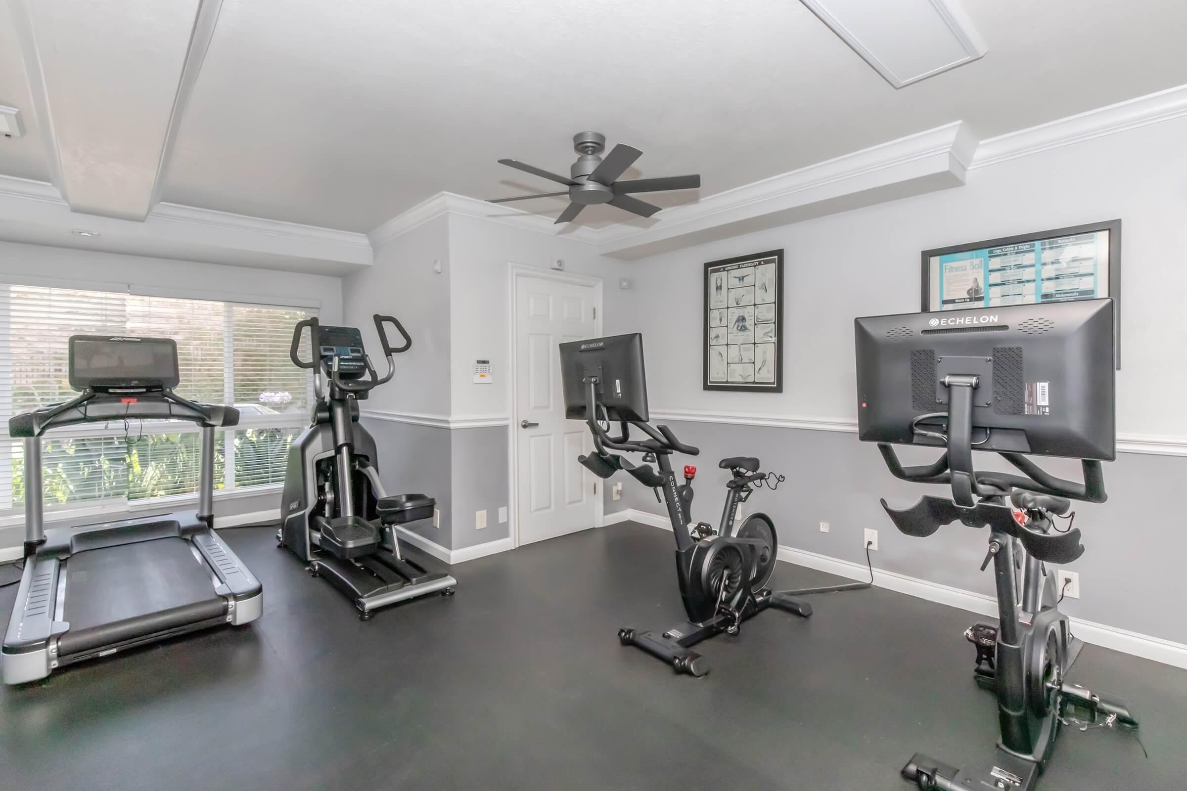 A modern home gym setup featuring a treadmill, an elliptical machine, and a stationary bike. The room has a sleek black floor, a ceiling fan, and large windows allowing natural light. A wall displays framed fitness-related documents.