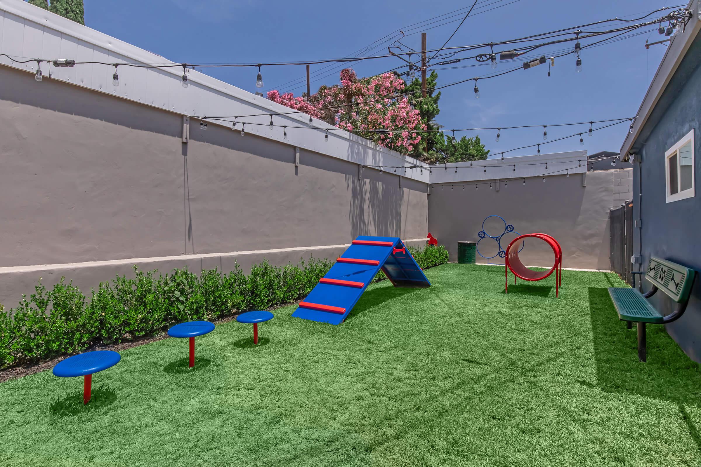 A colorful outdoor play area featuring a blue and red climbing structure, a circular tunnel, and several blue stools set on artificial grass. The space is bordered by low green shrubs and has string lights overhead, with pink flowering trees in the background under a clear blue sky.