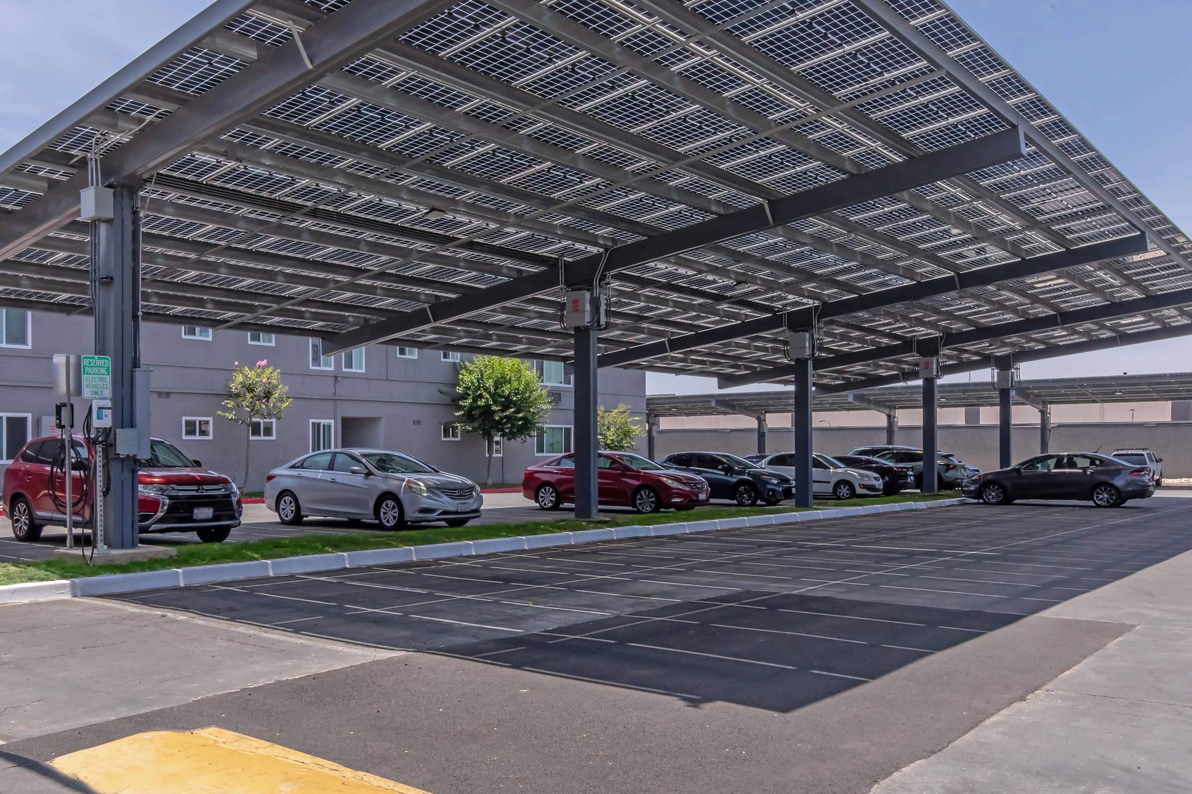 A shaded parking area with several cars parked under large solar panel canopies. The setting is urban, with a building visible in the background. The scene is clear and sunny, showing a sustainable approach to vehicle parking.