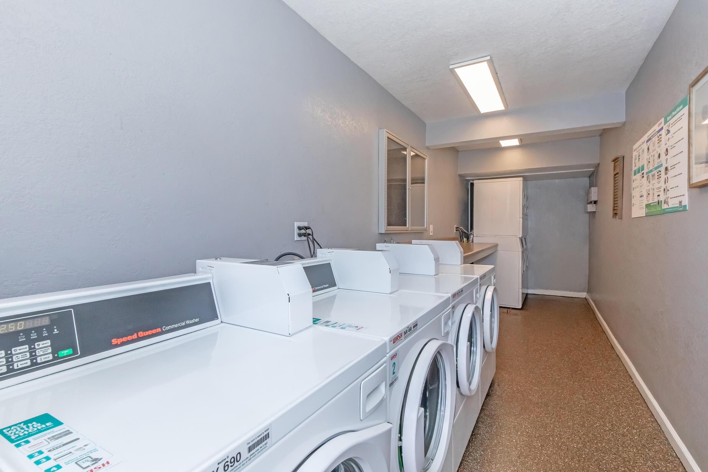 A laundry room featuring several white washing machines and dryers lined up against a wall. The space has gray walls, a door at the end, and overhead lighting. Laundry instructions and information are displayed on a nearby wall. The floor has a speckled pattern, and the area is well-lit and tidy.
