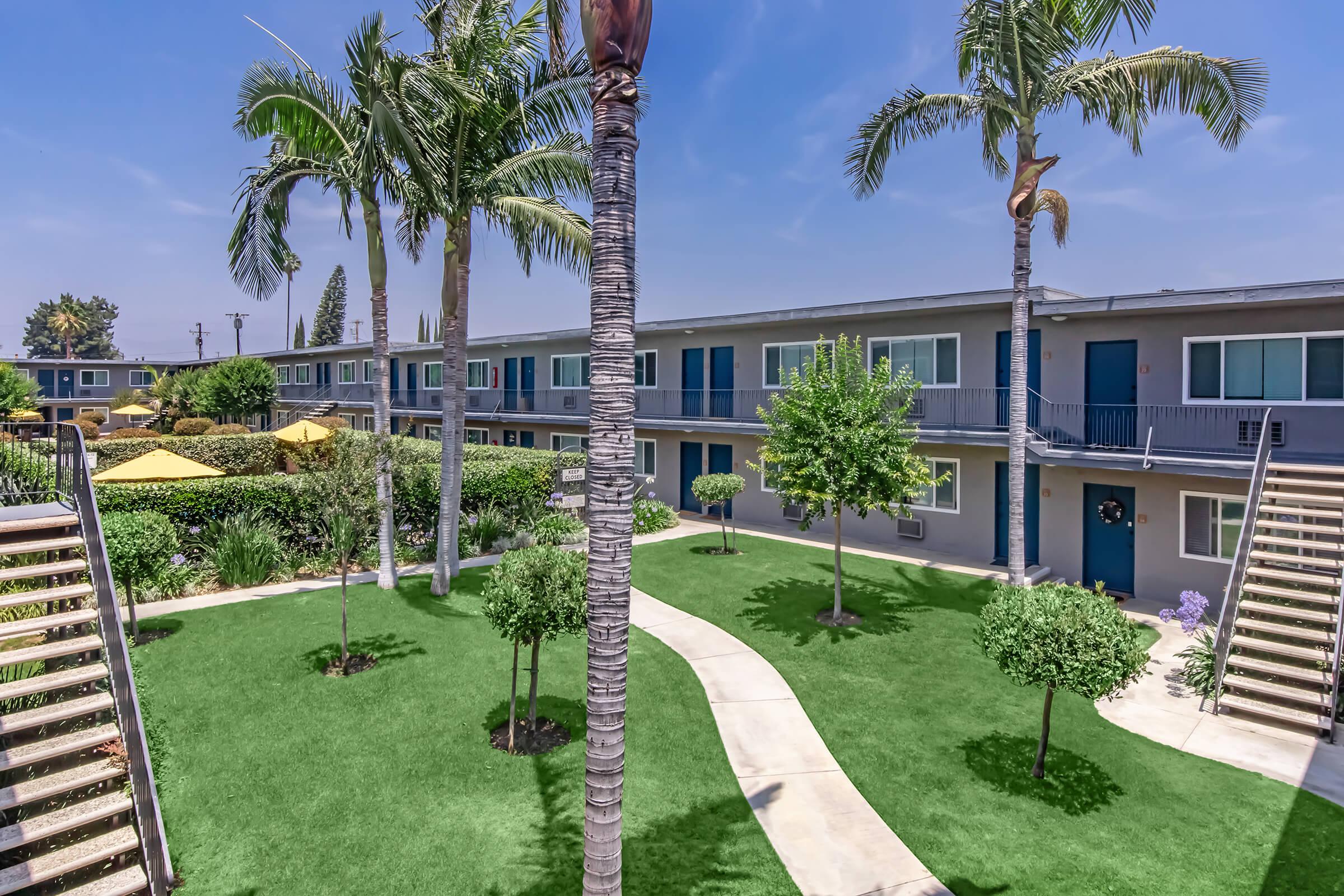 A landscaped courtyard featuring palm trees, well-maintained grass, and small shrubs. Surrounding the area are two-story apartment buildings with blue doors. The sky is clear and sunny, adding to the inviting atmosphere of the community.